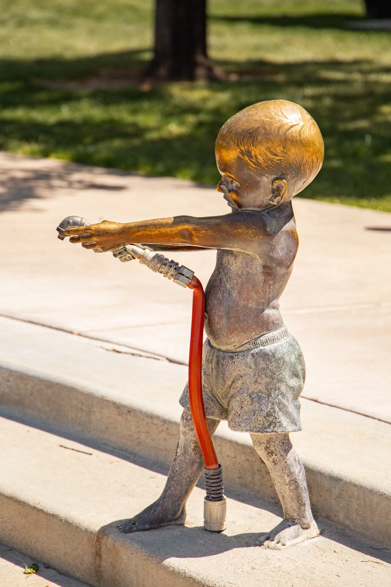 A bronze sculpture of a child holding a hose, with a dog standing on its hind legs touching the child's hand, set outdoors on a concrete sidewalk with grass and trees in the background.