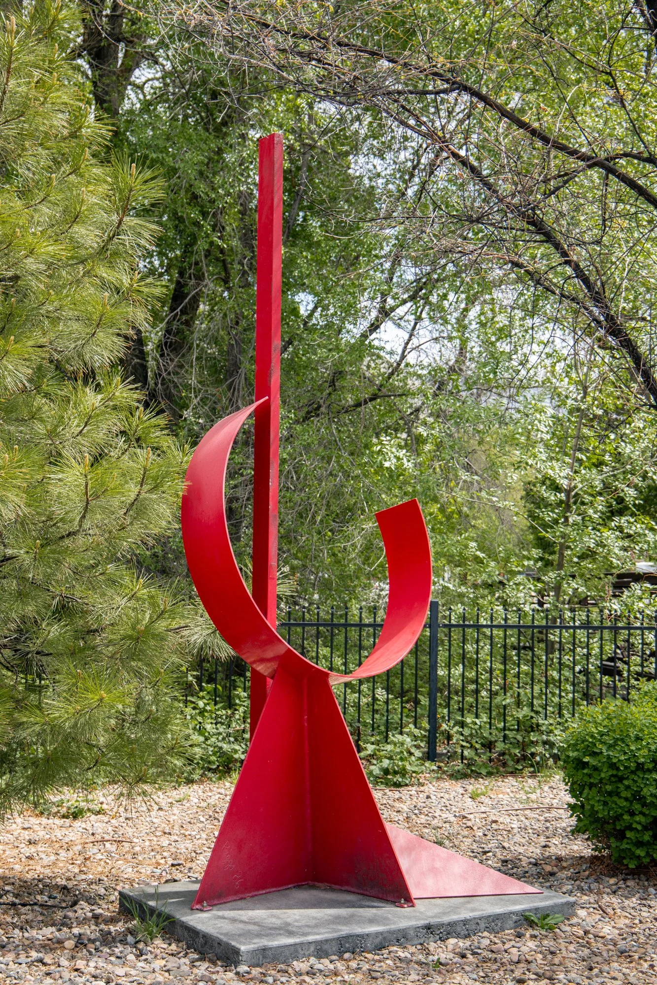 A modern abstract red sculpture installed outdoors on a concrete base, surrounded by trees and greenery.