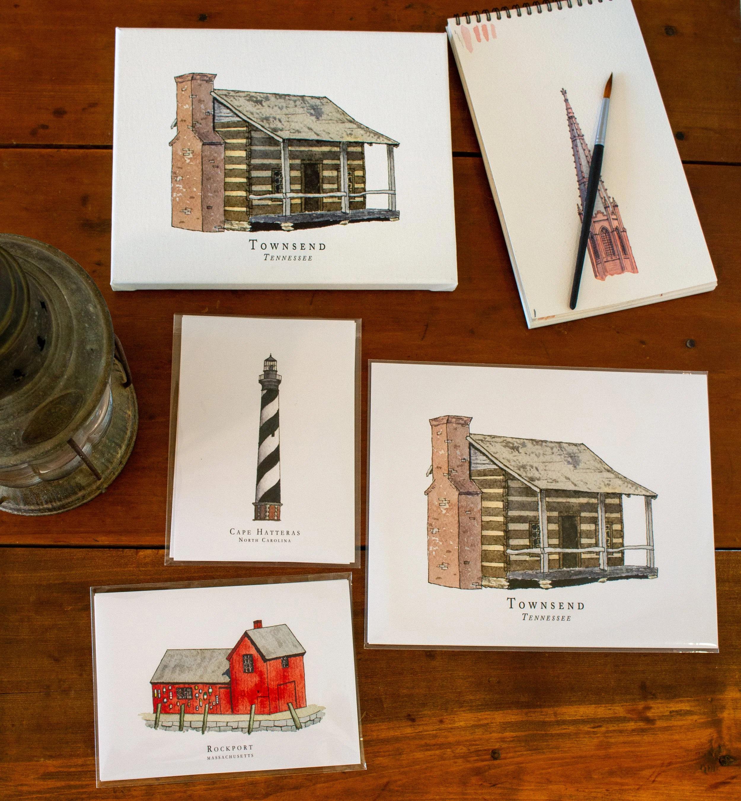 Postcards featuring watercolor illustrations of historic lighthouses and buildings from Townsend, Tennessee, Cape Hatteras, North Carolina, and Rockport, Massachusetts, displayed on a wooden surface.