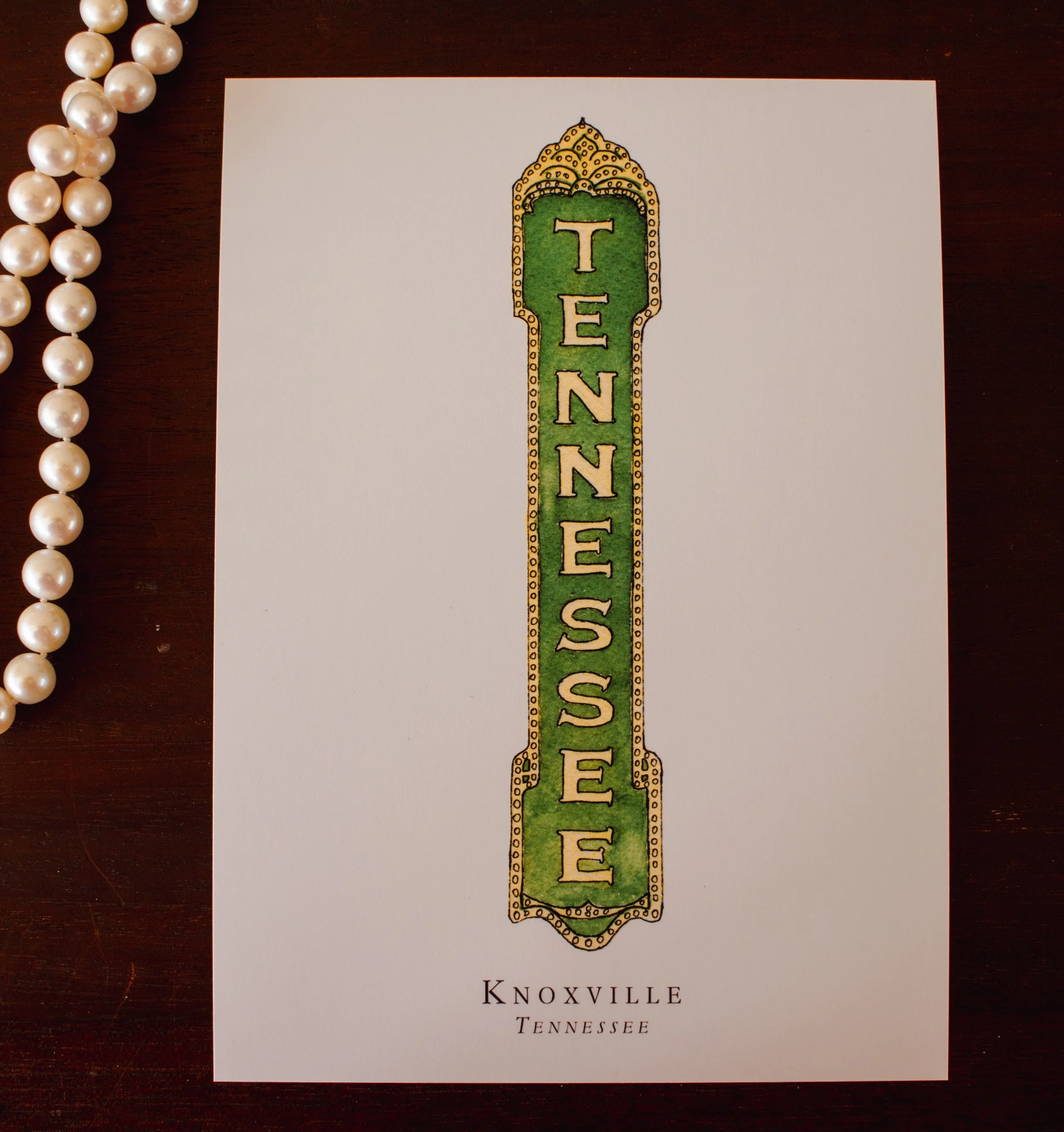 A white paper with the word "Knoxville" written at the bottom, a green and yellow train-shaped design in the center with the word "Tennessees" vertically inside it, and a pearl necklace on the dark wooden surface beside it.