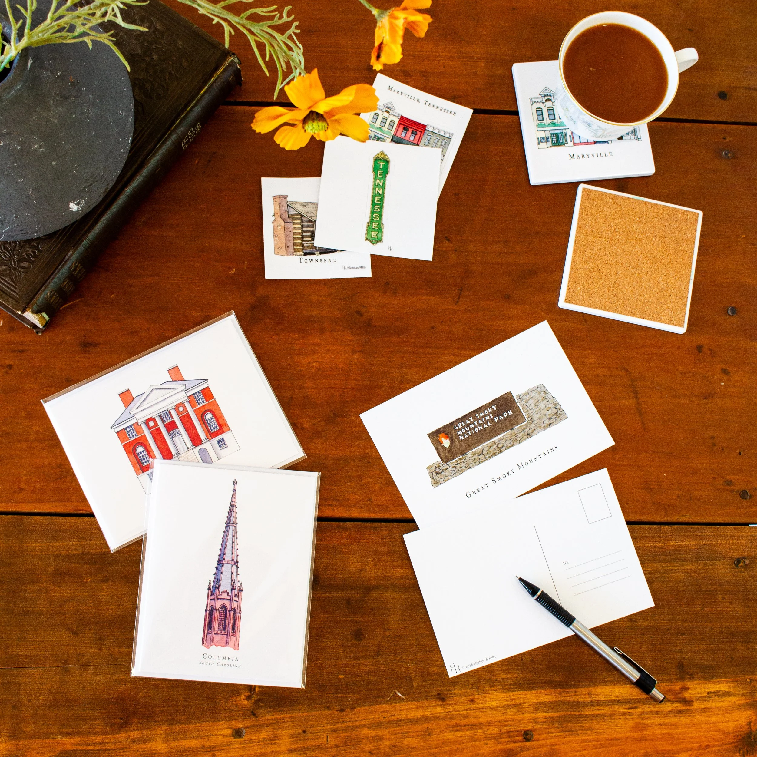 Top-down view of a wooden table with illustrated cards of famous landmarks, a cup of coffee on a coaster, a small plant in a black pot, a dark hardcover book, and a pen.