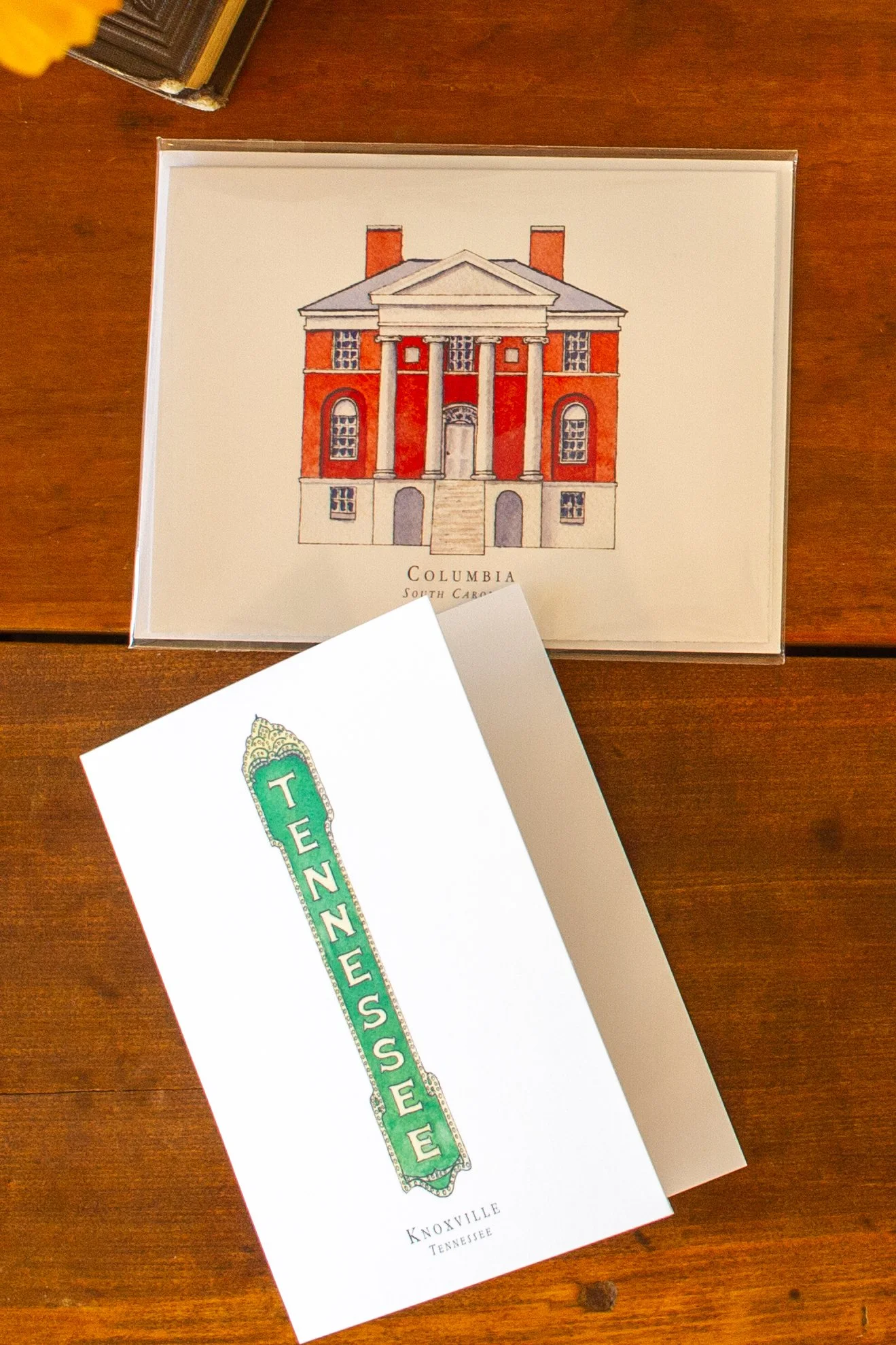 Illustration of a large red brick house with white columns and a staircase, labeled 'Columbia, South Carolina.' In the foreground, there's a white sheet with a green and white sign that reads 'TEEZEUS MUSEUM,' and a small part of a booklet or paper showing the words 'Knoxville Tennessee.'