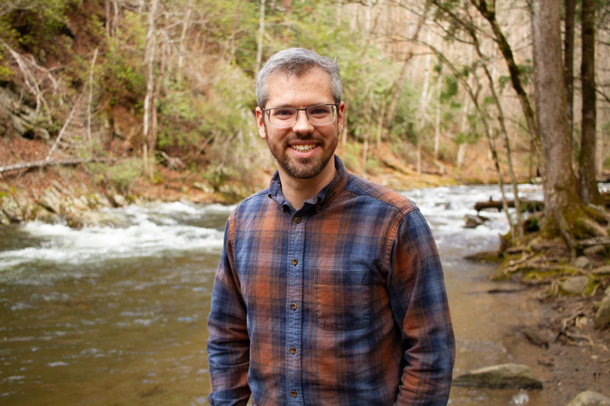 A smiling man with glasses and a beard standing outdoors near a stream in a forest with leafless trees and some green foliage.