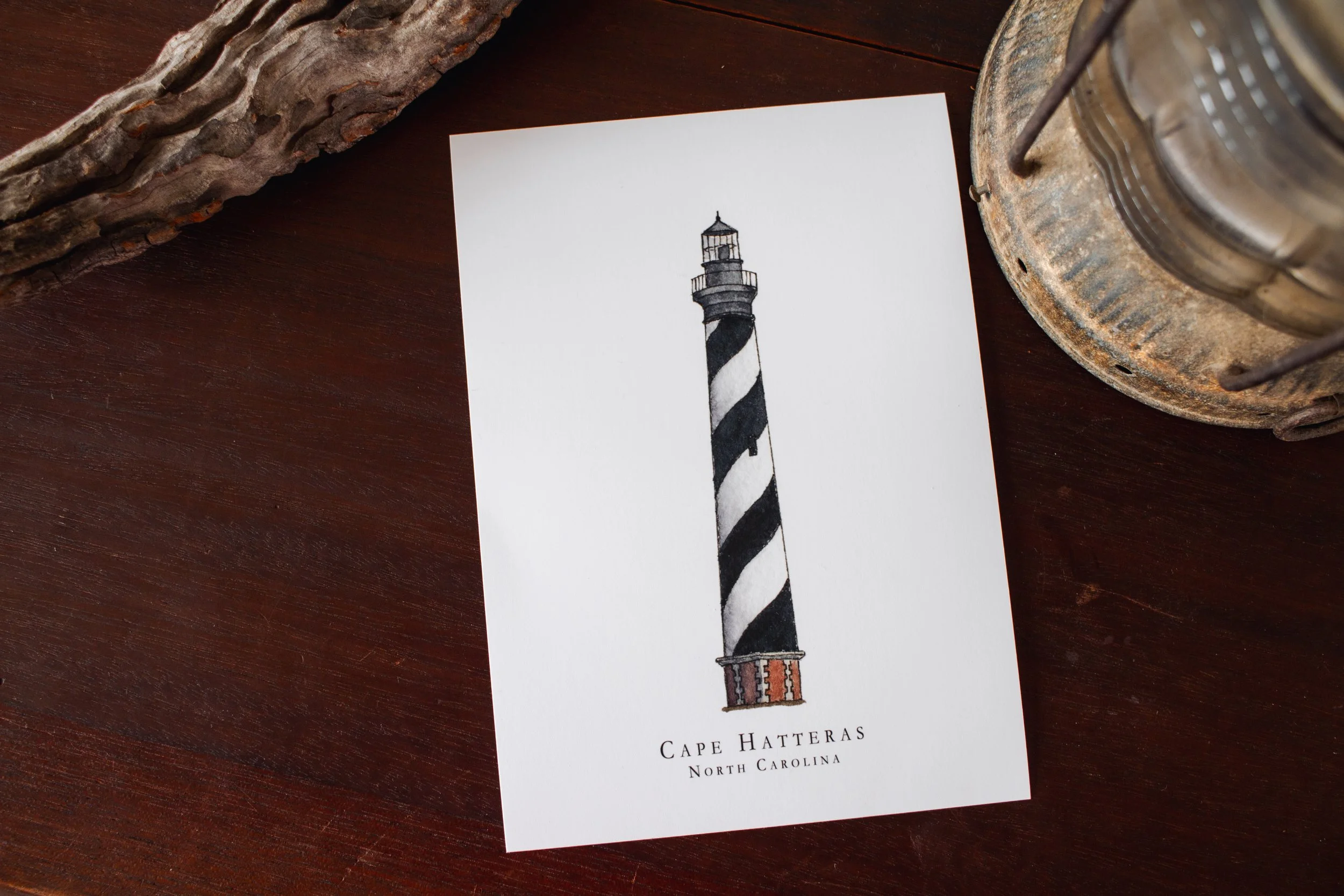A printed illustration of the Cape Hatteras lighthouse in North Carolina on a piece of white paper, placed on a dark wooden table surrounded by natural wood and metal decorative objects.