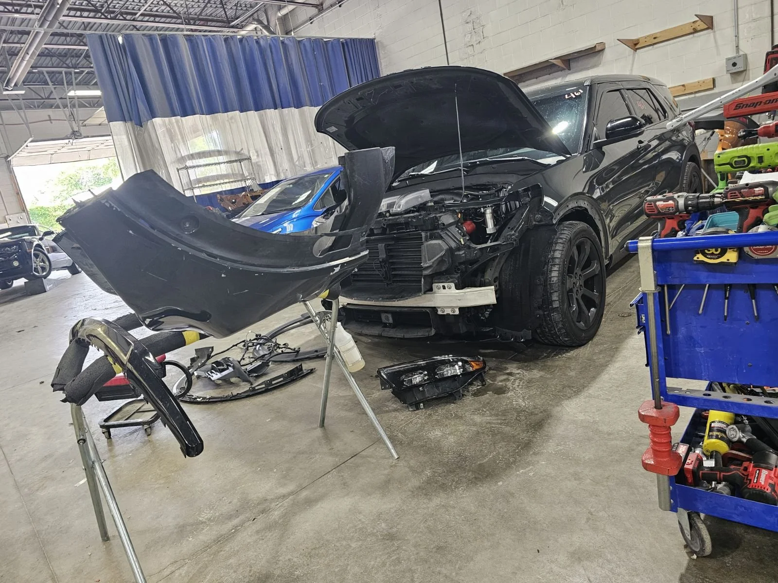 Interior of a car repair shop with a black SUV undergoing repair, its front bumper and parts disassembled and on a stand. Tools and parts are scattered around, and a blue cart with tools is visible on the right.