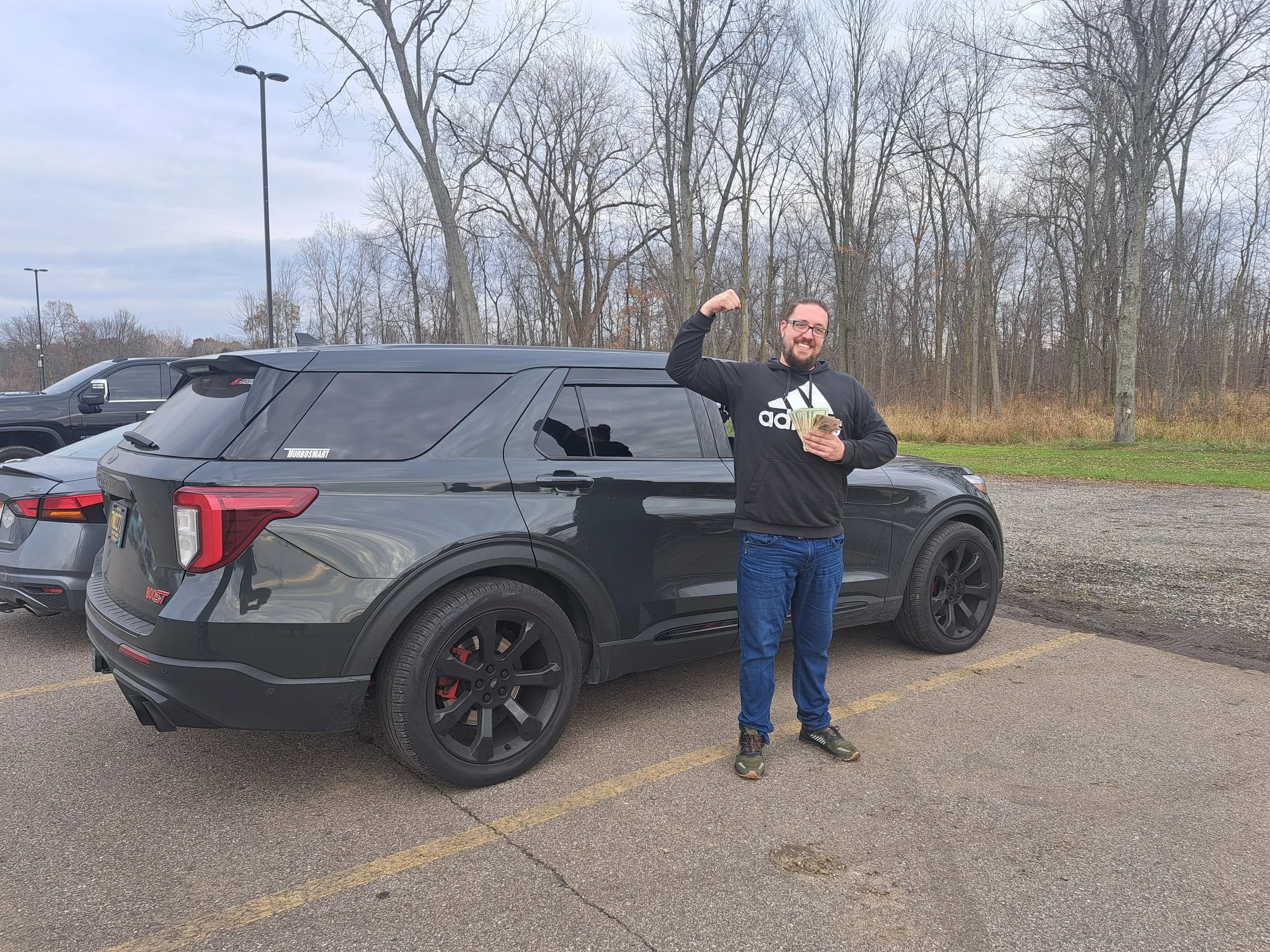 A man standing in a parking lot next to a black SUV, holding cash in one hand and flexing his bicep with the other, smiling.