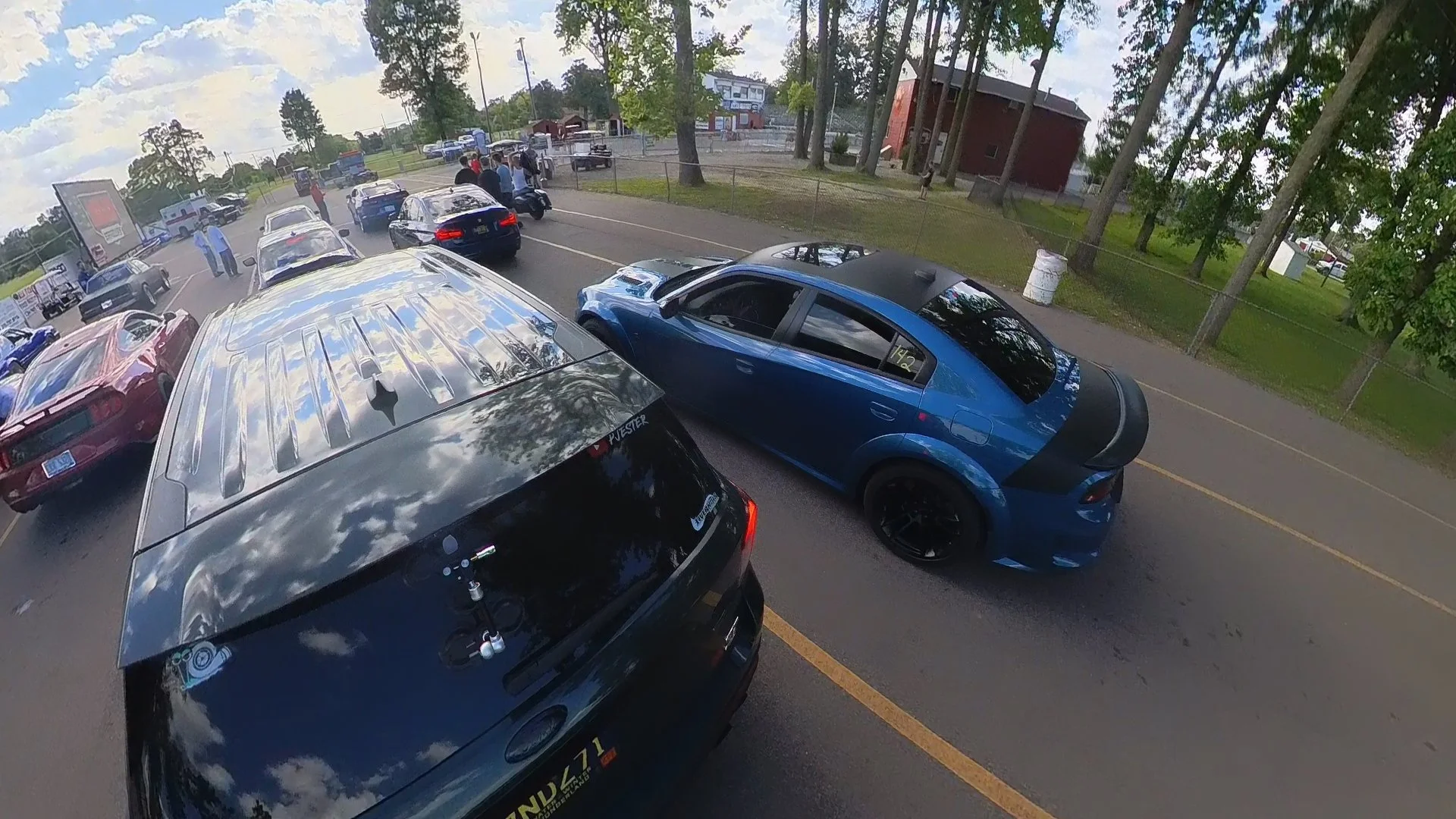 Drag strip staging lanes filled with various cars, with a blue Charger Hellcat in the foreground and other vehicles and people in the background. Trees and a small building are visible beyond the parking lot.