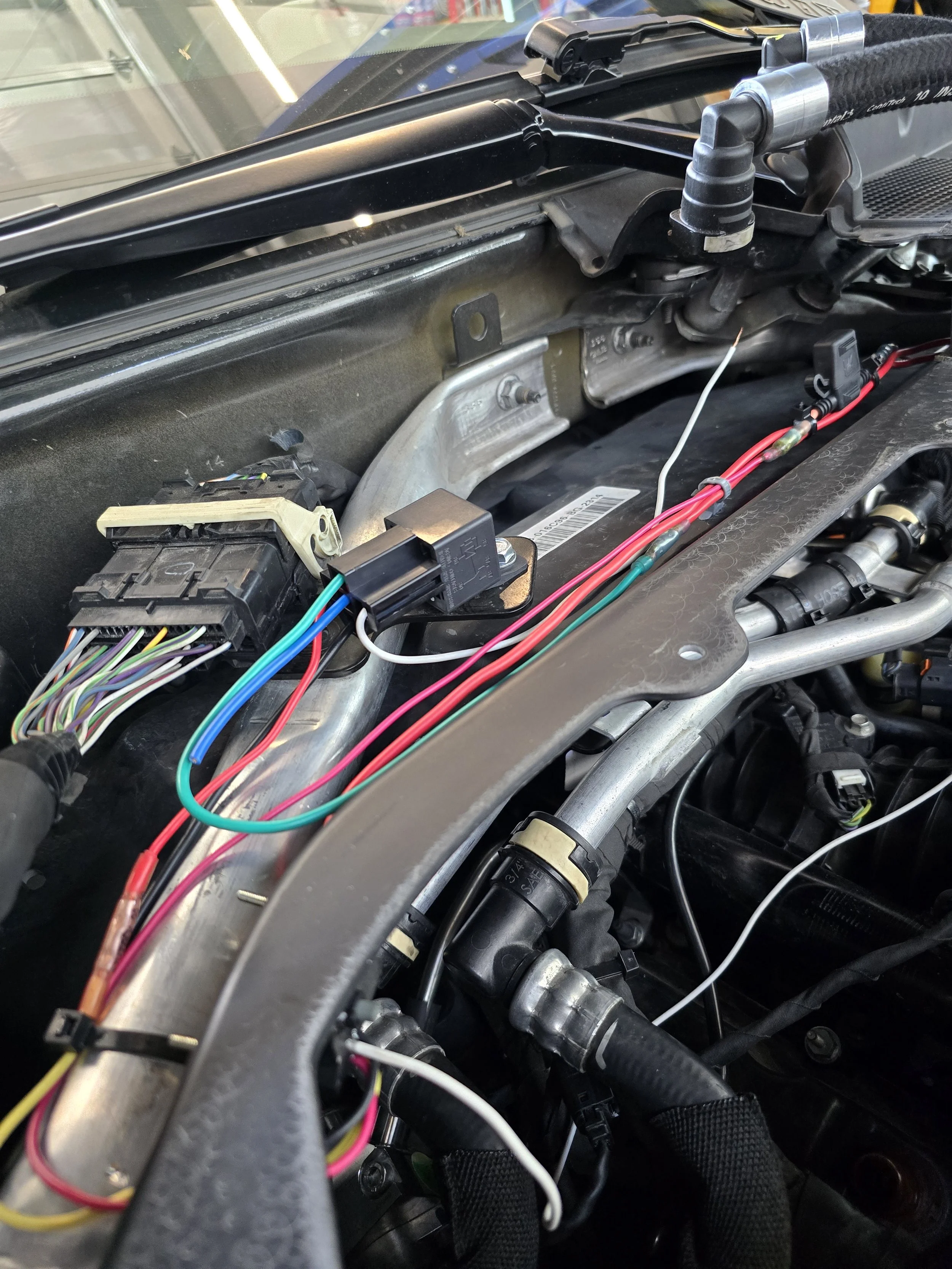 Car engine bay with exposed wiring, connectors, and mechanical parts, indicating ongoing maintenance or repair.