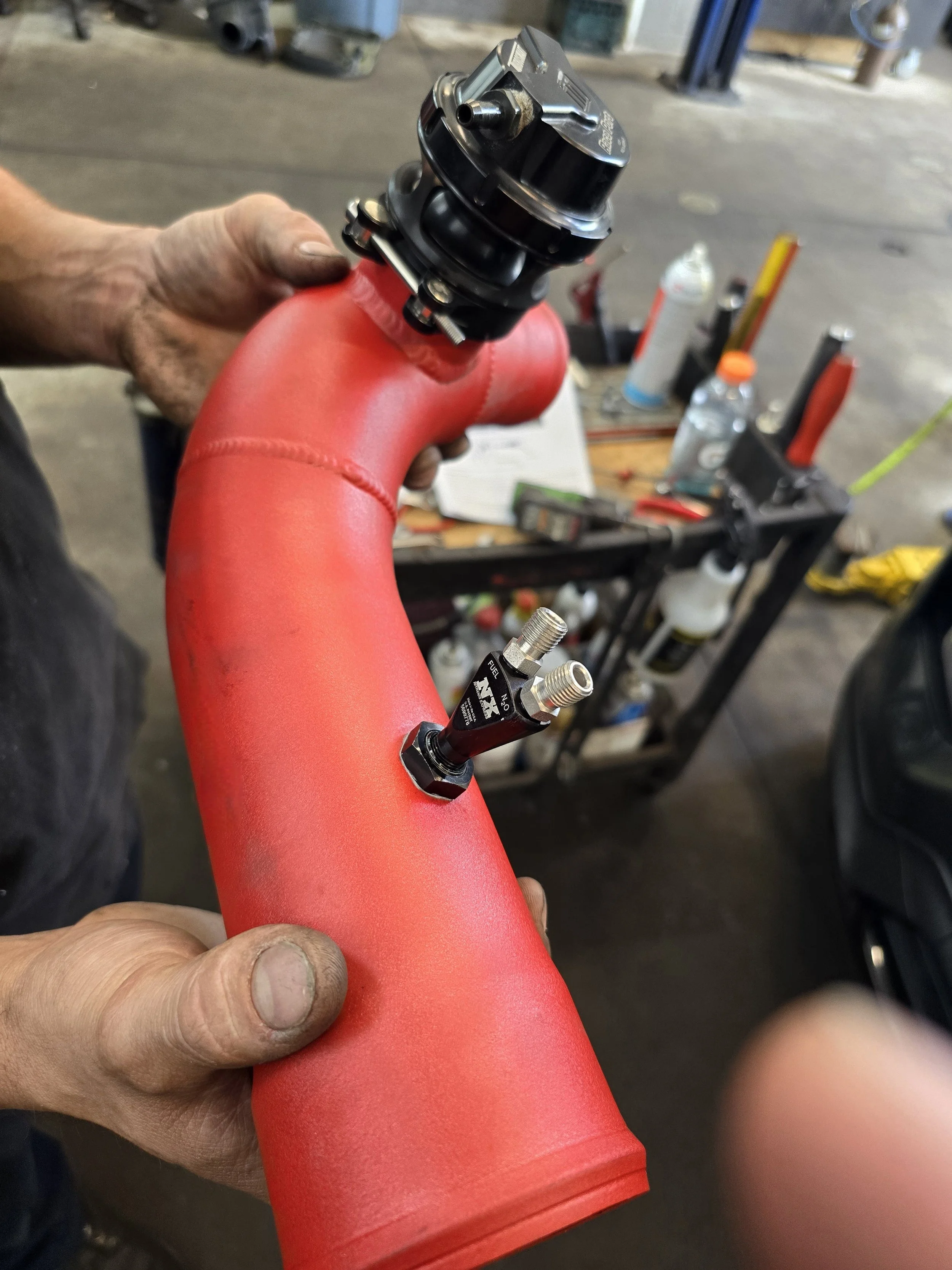 A person holding an aluminum charge pipe with metal connectors and a black nitrous splitter in a garage or workshop setting.