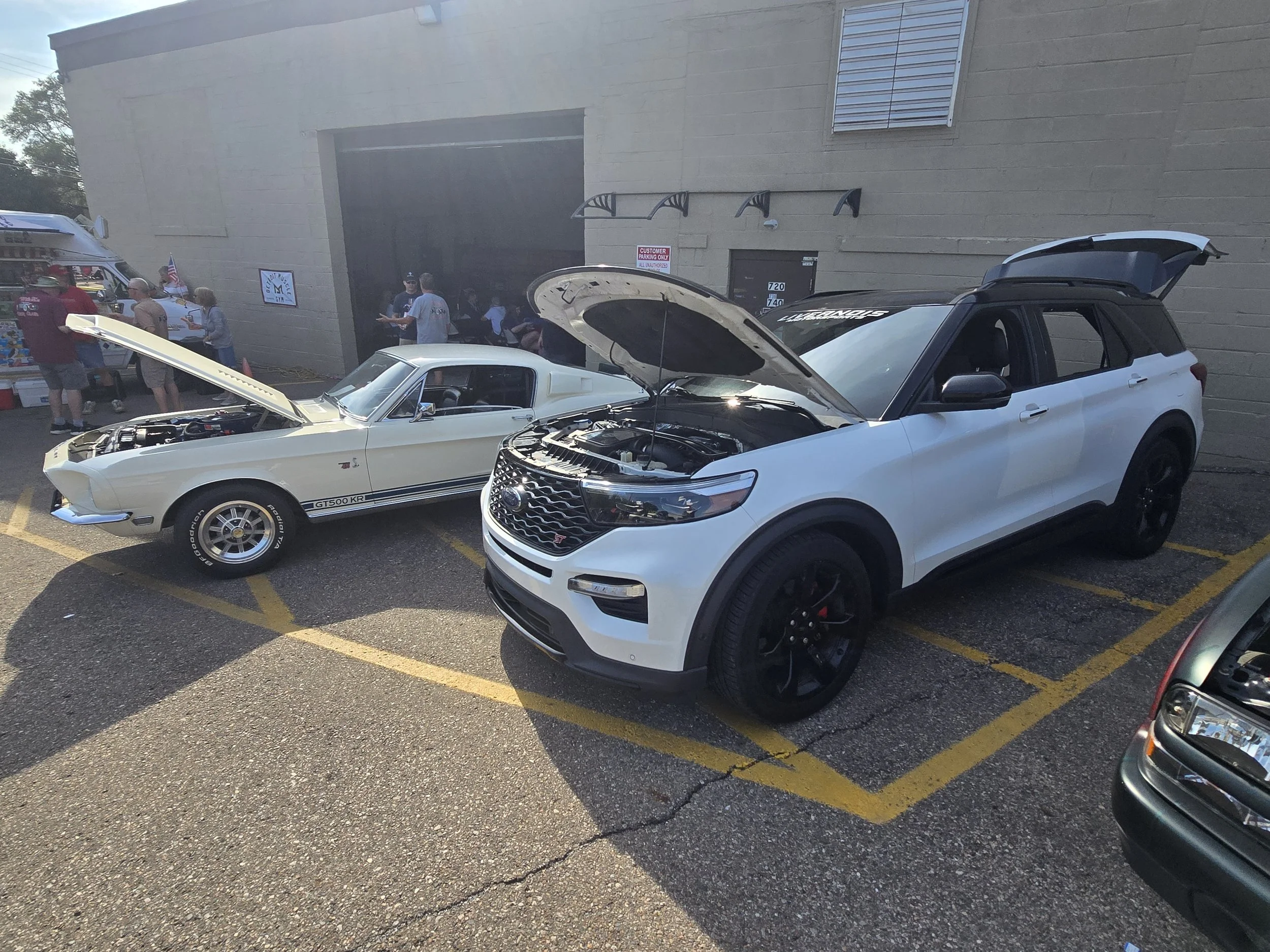 A Ford Explorer ST and a vintage white muscle car with opened hoods on display at a car show.