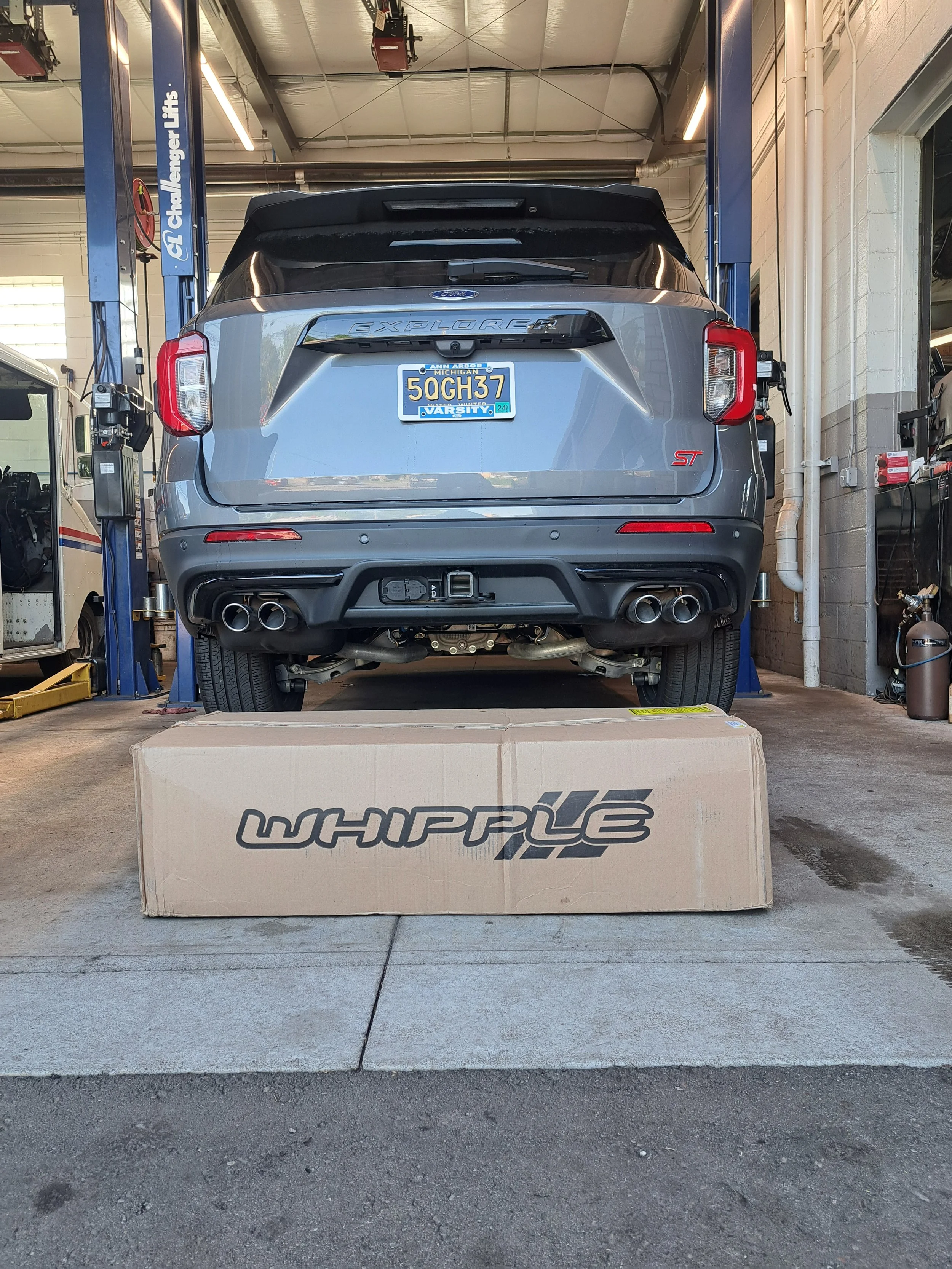 A gray Ford Explorer ST SUV inside a vehicle repair shop, positioned on a lift, with a Whipple Intercooler box placed on the ground in front of it.