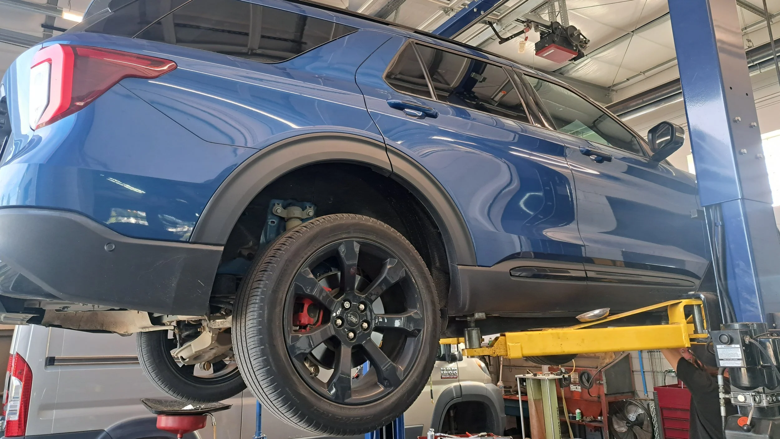 A blue SUV is lifted on a hydraulic lift inside an auto repair shop. The rear left tire is removed, exposing the brake assembly.