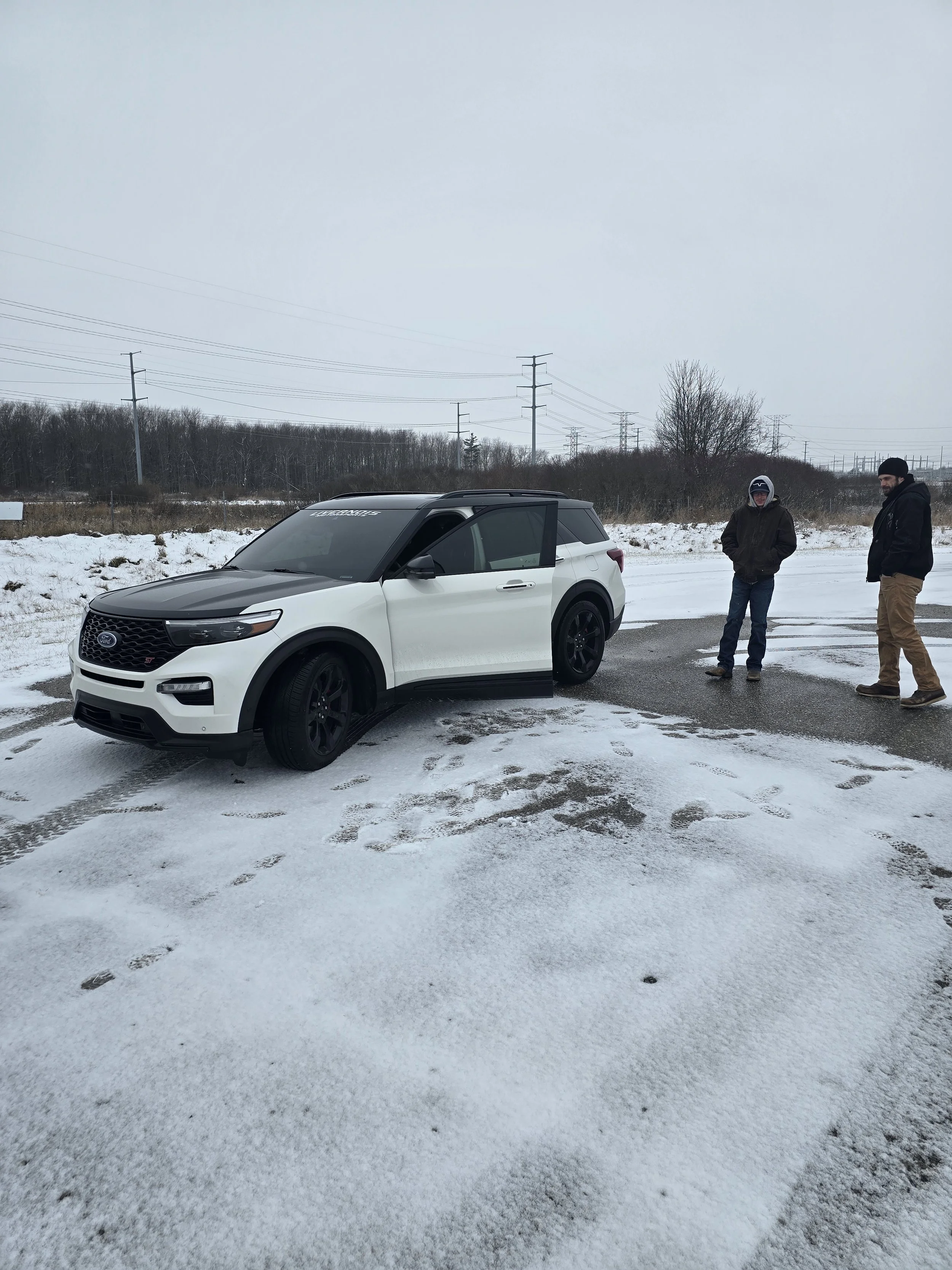 A white and black SUV parked on a snowy road with two men standing nearby, wearing winter clothing, in a cold, overcast outdoor setting.