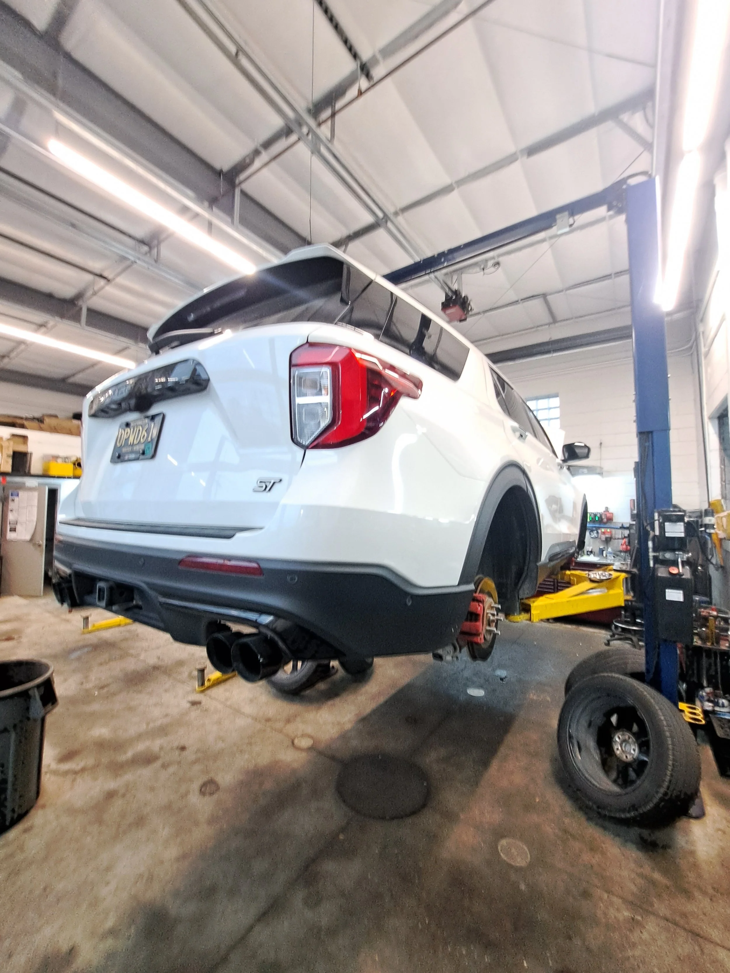 A white SUV, elevated on a hydraulic lift in an auto repair shop, with removed wheels and visible exhaust pipes.