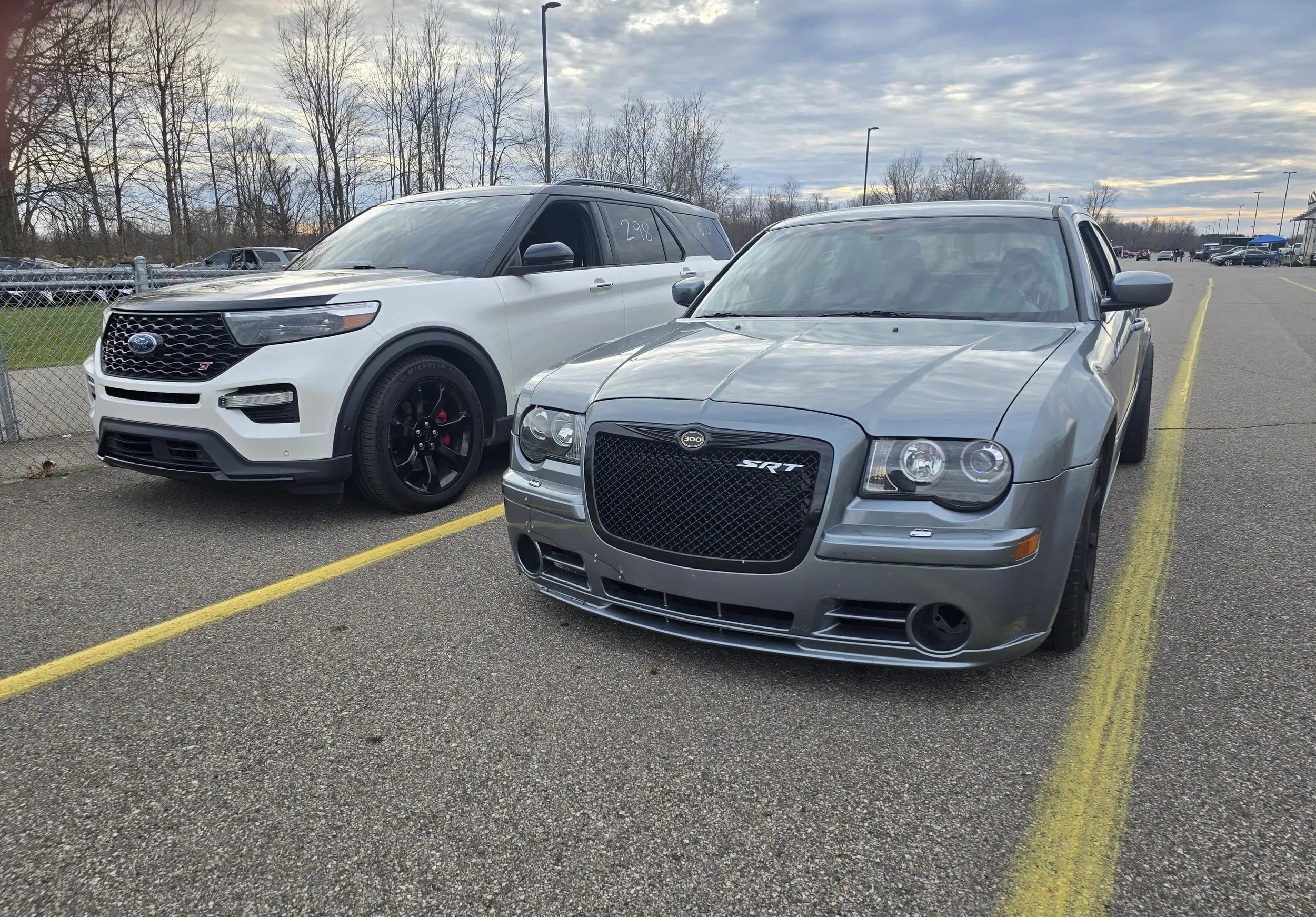 A silver Dodge Charger SRT and a white Ford Explorer parked at a drag strip with other vehicles and a cloudy sky in the background.