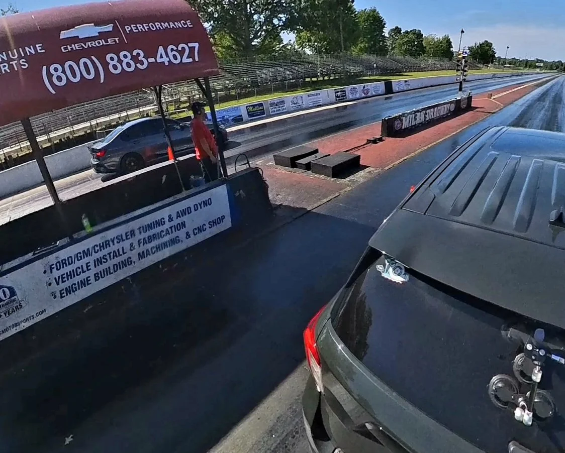 Drag racing starting line with a car on the starting line and a person standing behind the rail. The drag strip has advertising banners and a red starting light tree, with a vehicle in the foreground.