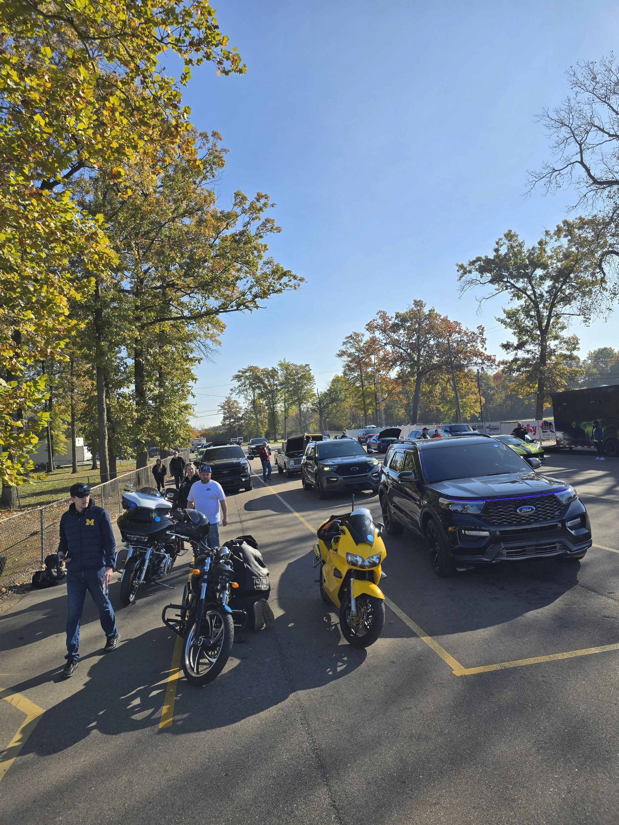 A parking lot with several motorcycles and cars, with a few people walking around on a bright, sunny day. There are trees with autumn leaves in the background.