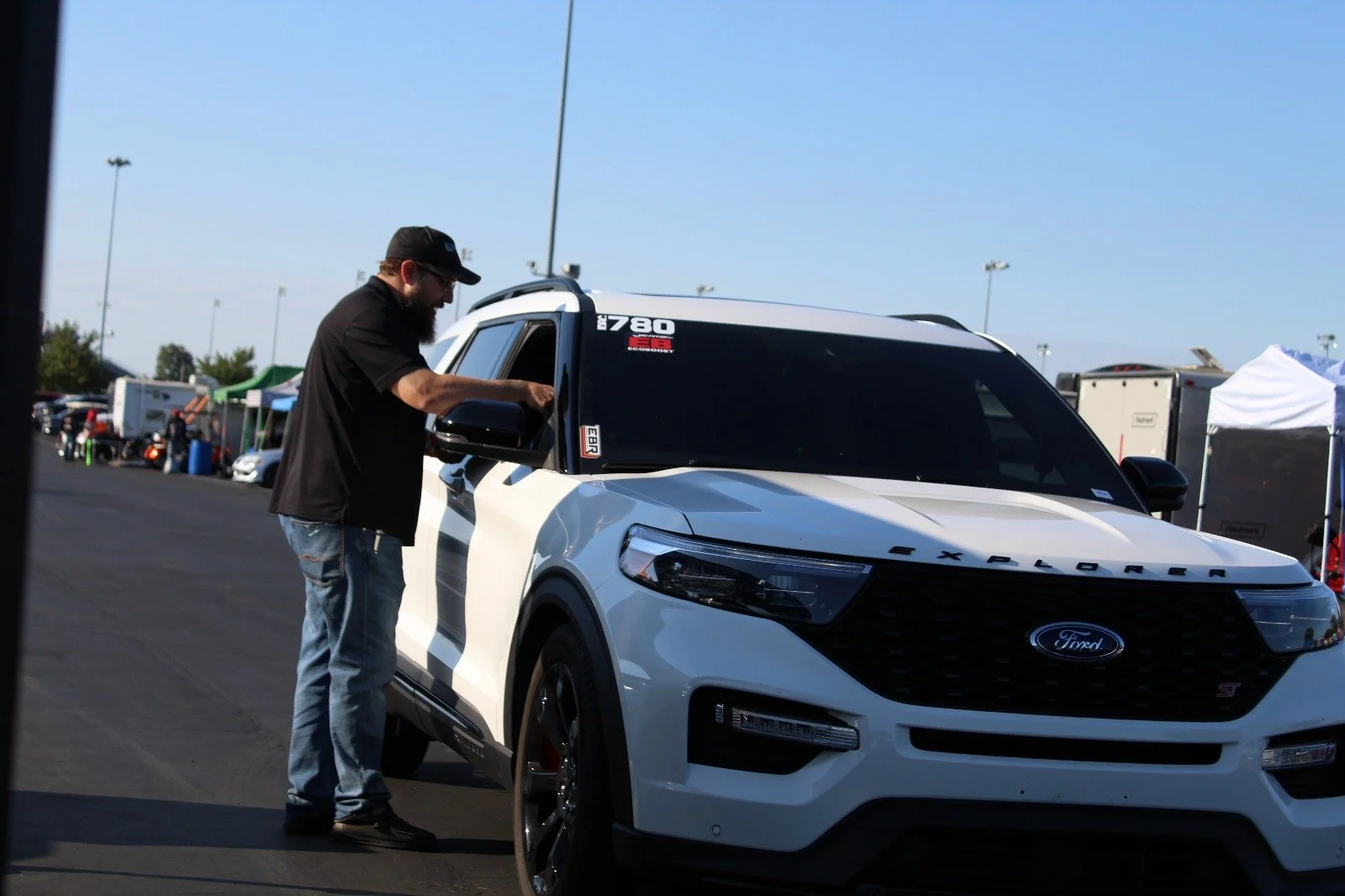 Pjester wearing a black shirt, jeans, and a cap stands next to a white Ford Explorer parked at a race track or event lot, talking to the driver through the window, with tents and trailers visible in the background.