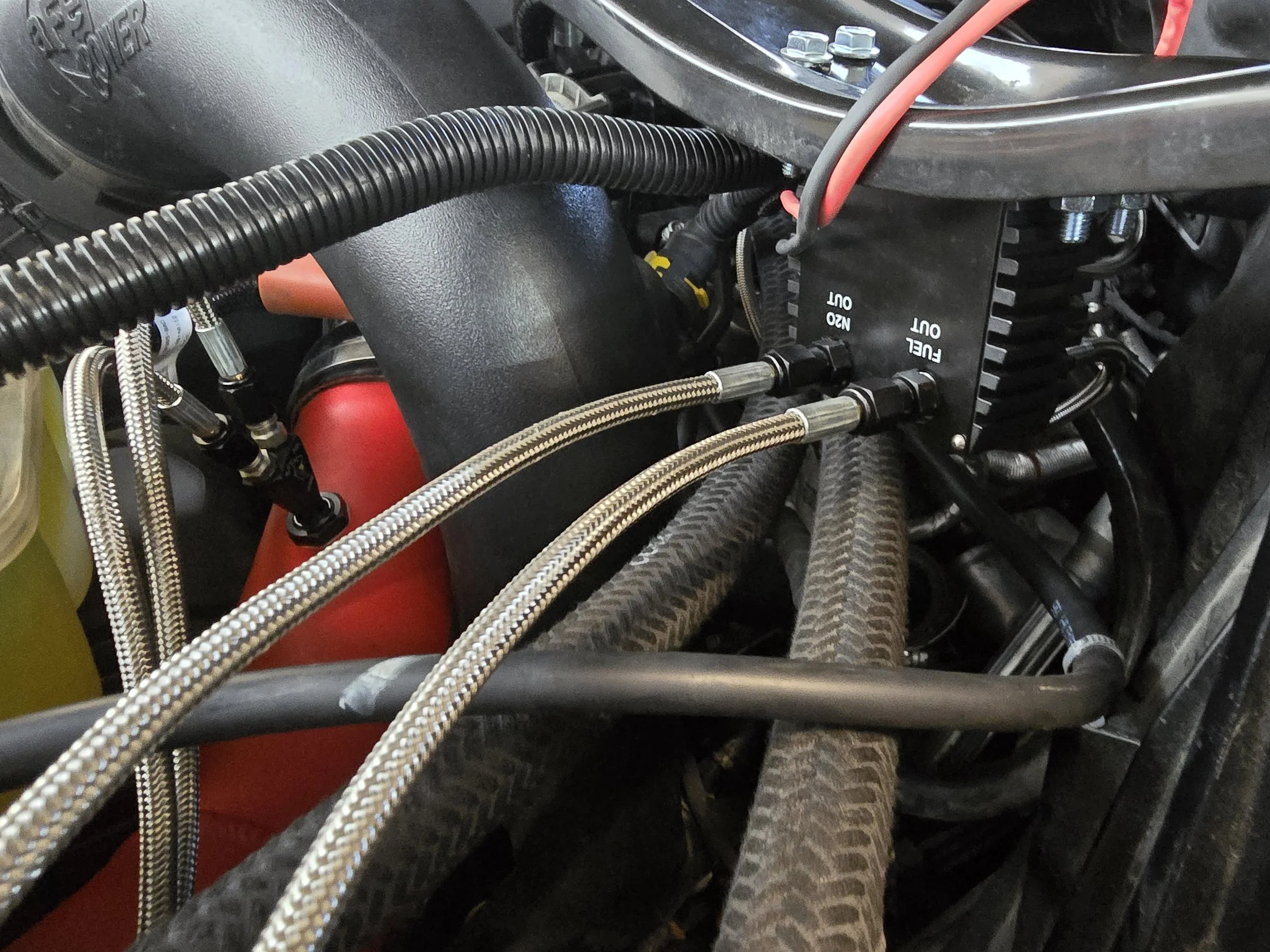 Close-up of a mechanical engine component with braided metal hoses, electronic wiring, and a black heat-resistant cover with labeled ports.