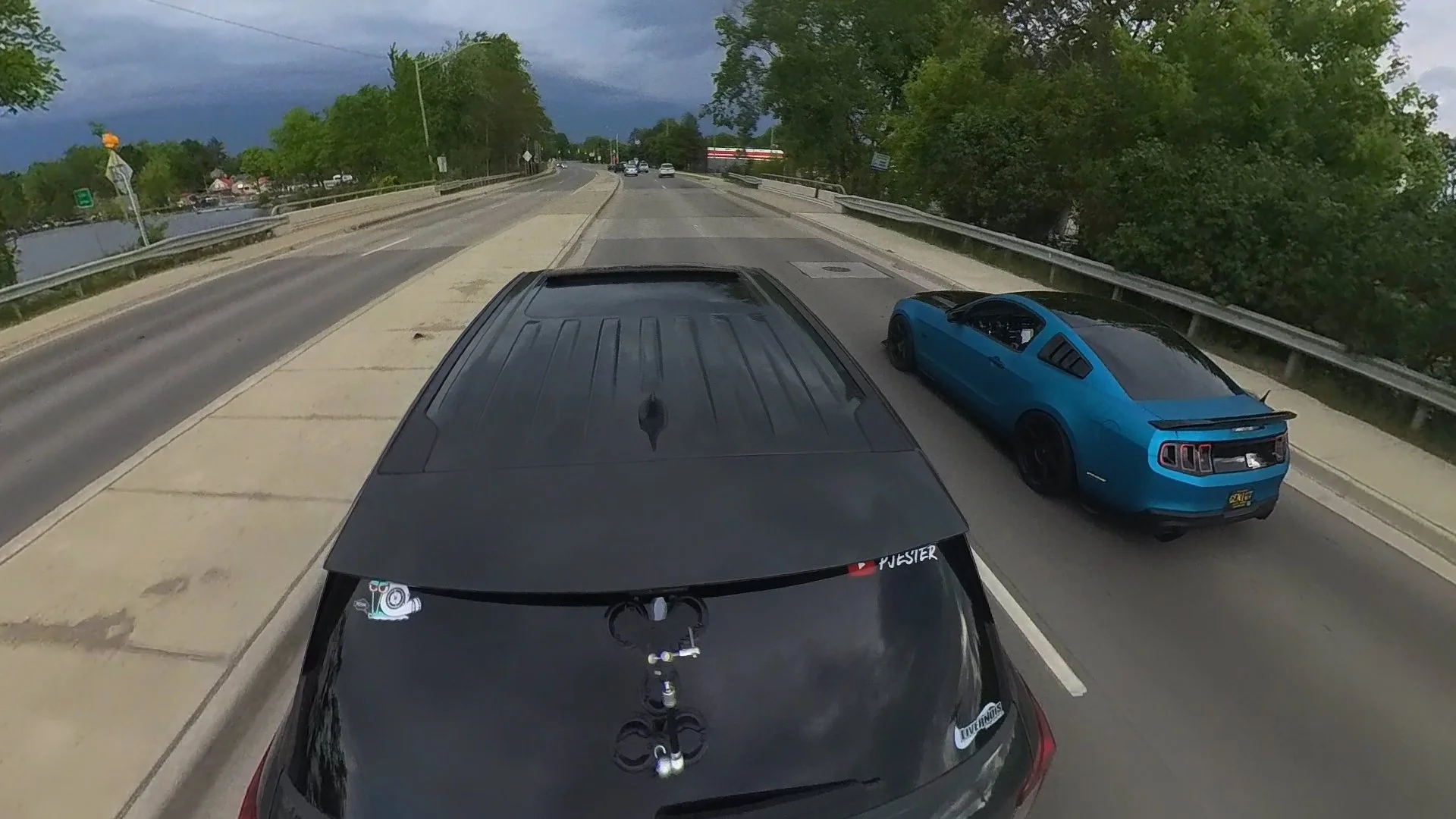 A black vehicle is on a two-lane road with a blue sports car to the right. The scene is outdoors with trees and a cloudy sky.