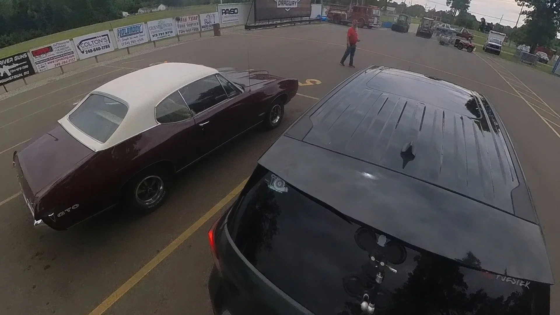A parking lot with a vintage maroon and white car and a modern black SUV parked side by side. A man in a red shirt is walking in the background, and there are advertisements on the fence along the perimeter.