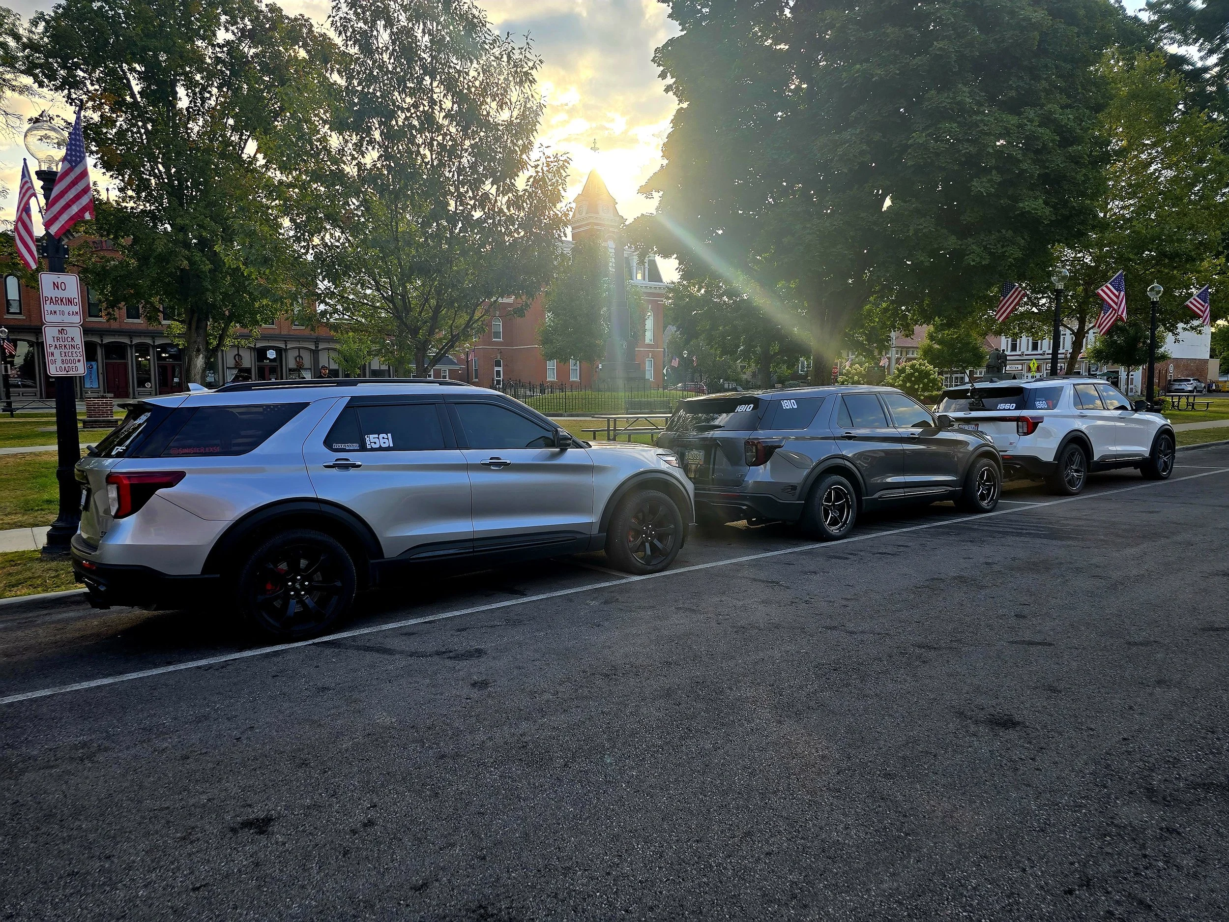 Three parked SUVs along a street with American flags on streetlamps, trees, and a historic building in the background, with sunlight filtering through the trees.