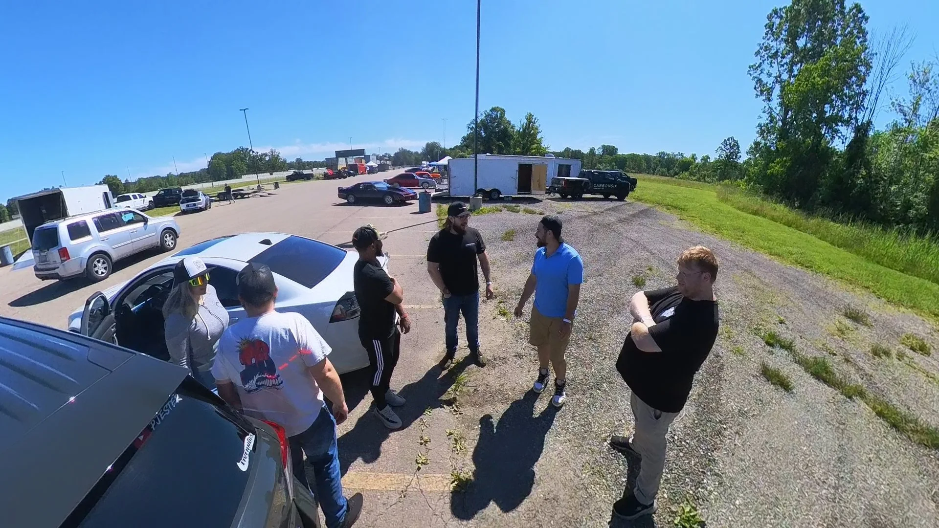 A group of seven people attending an Explorer ST event standing and talking in a parking lot on a sunny day, with several cars and trailers in the background and a grassy area with trees to the right.
