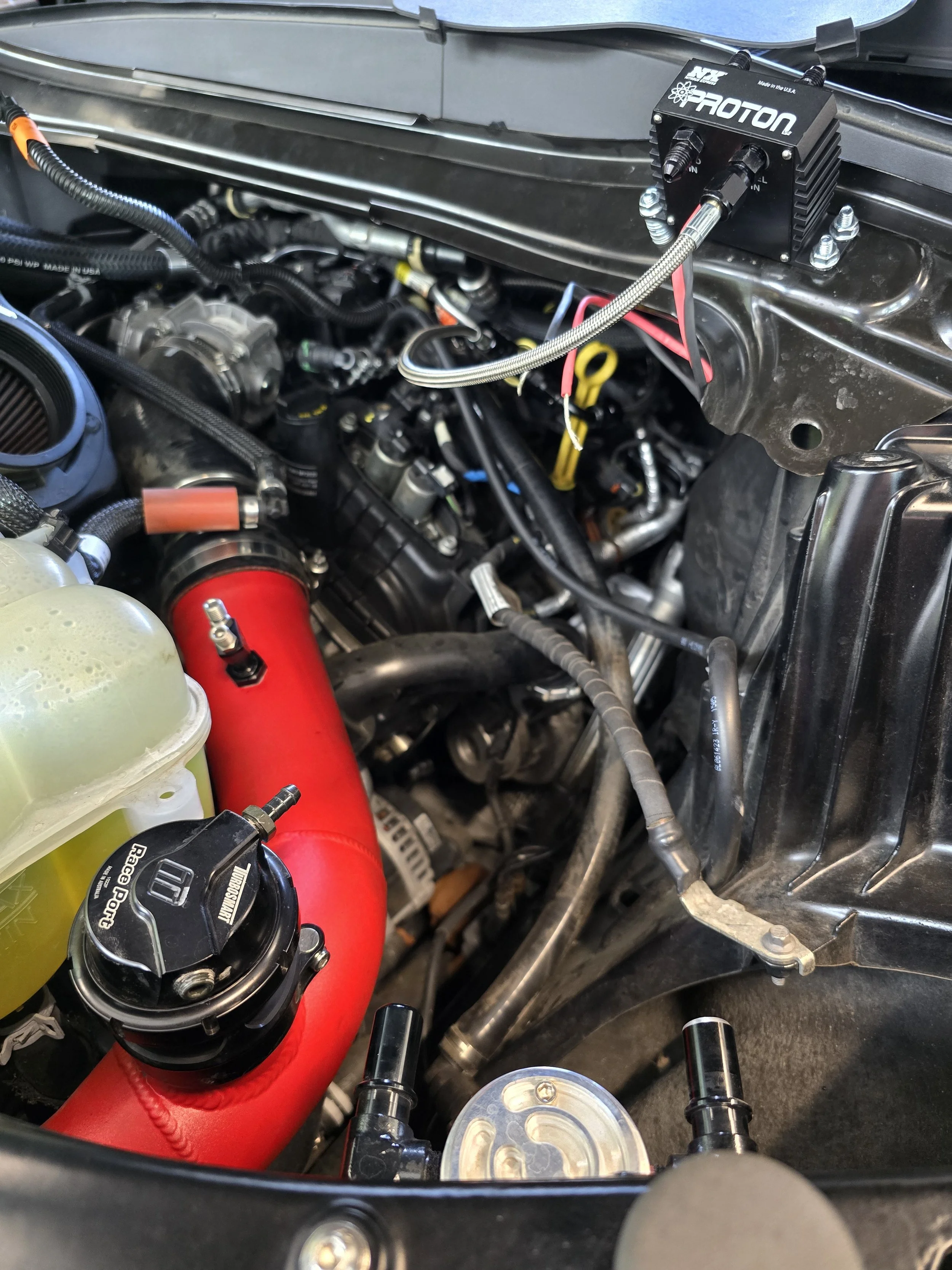 Close-up of a car engine bay with a red silicone hose, a yellow coolant reservoir, and a black Protron nitrous device mounted on the firewall.