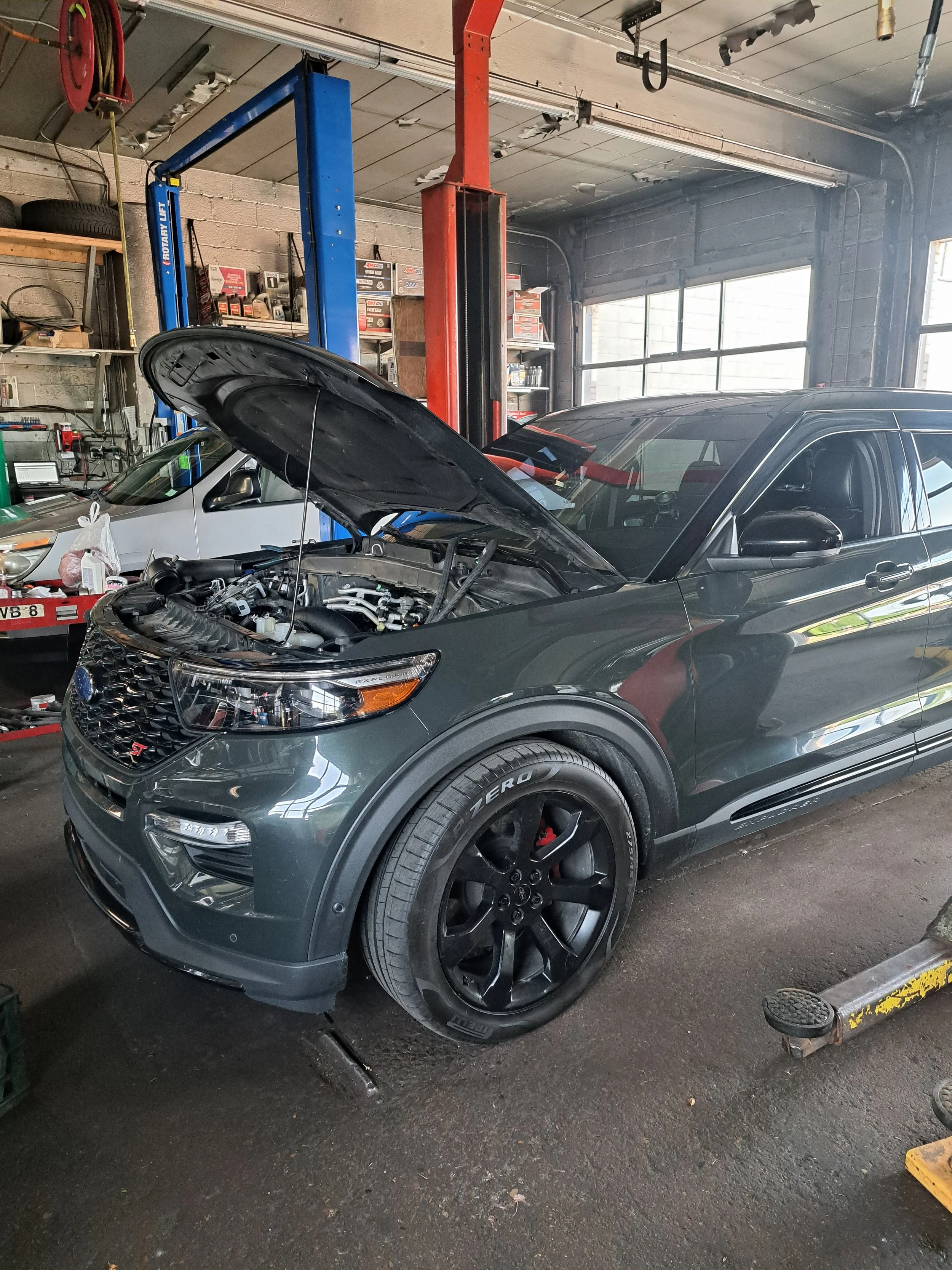 A gray SUV with its hood open inside an auto repair shop, with tools and equipment in the background.