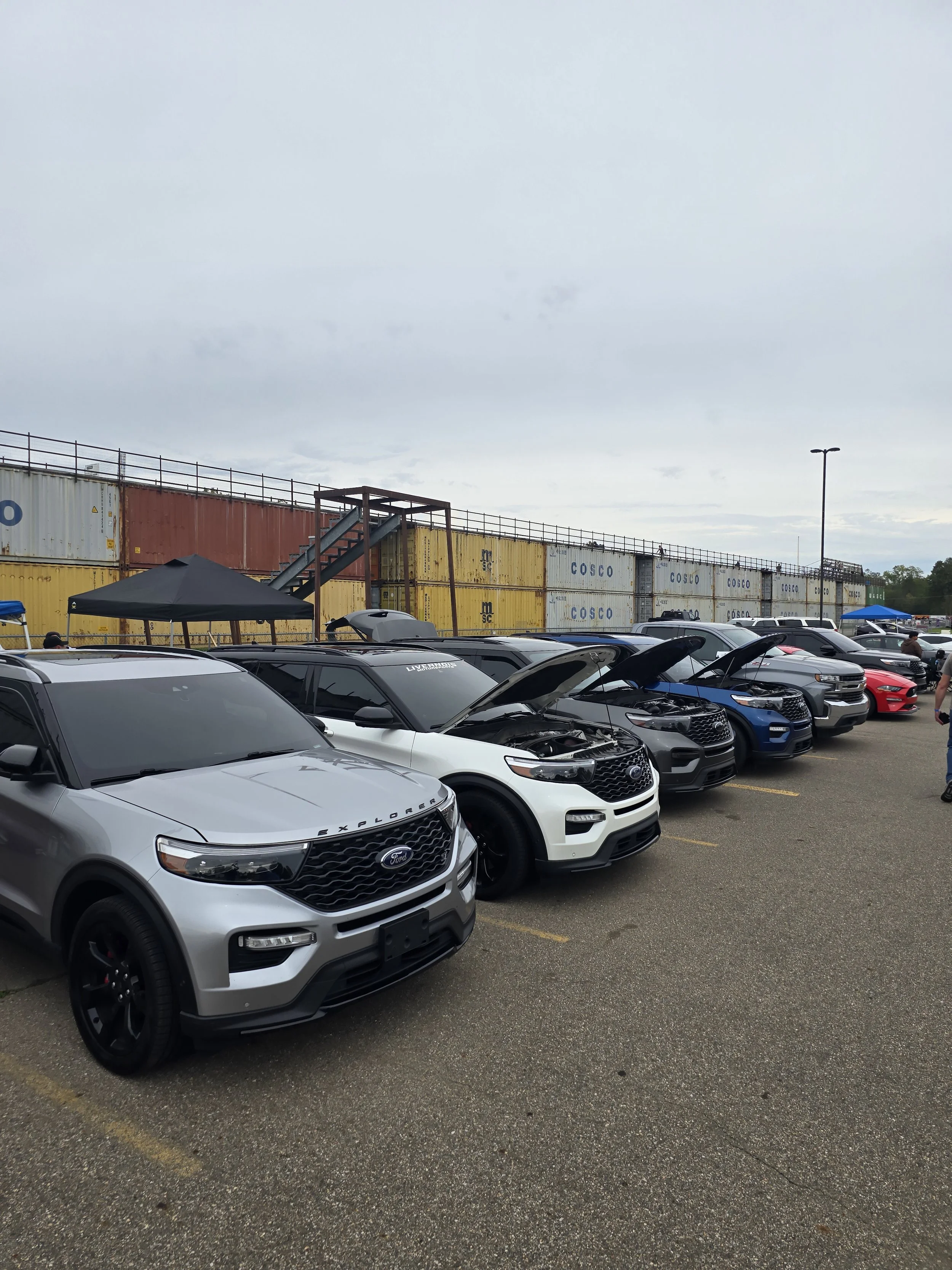 Multiple cars parked at a drag strip next to stacked cargo containers under a cloudy sky.