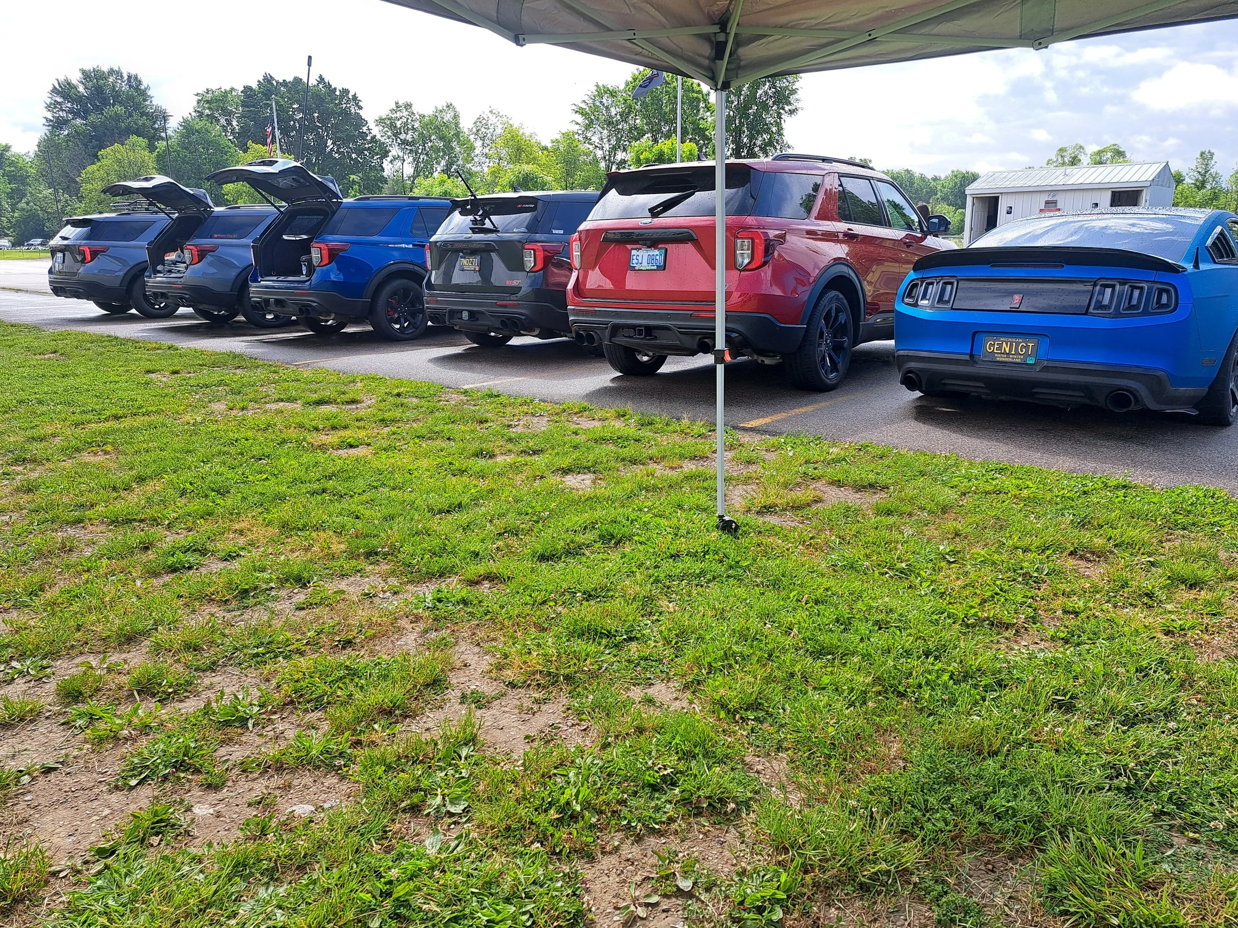 A row of six parked SUVs, with some having open tailgates, under a canopy in a parking lot on a partly cloudy day, with grass in the foreground and a small white building in the background.