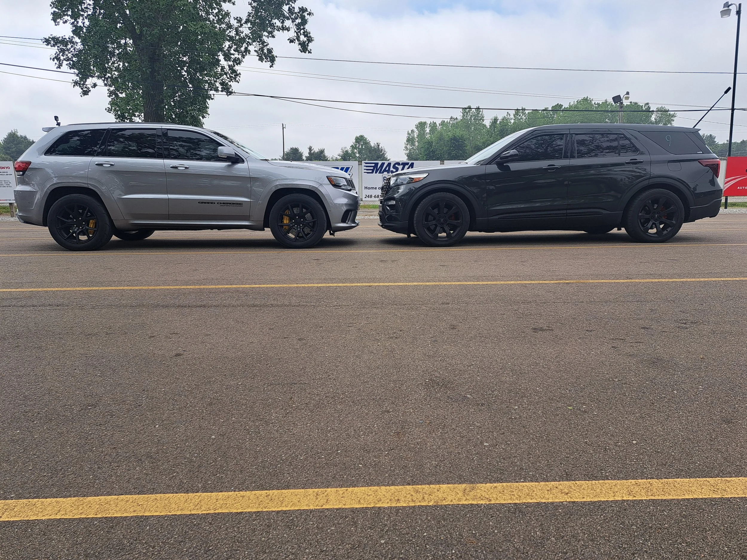 Two SUVs parked on a street facing each other, with trees and signs in the background. Jeep Trackhawk vs. Explorer ST