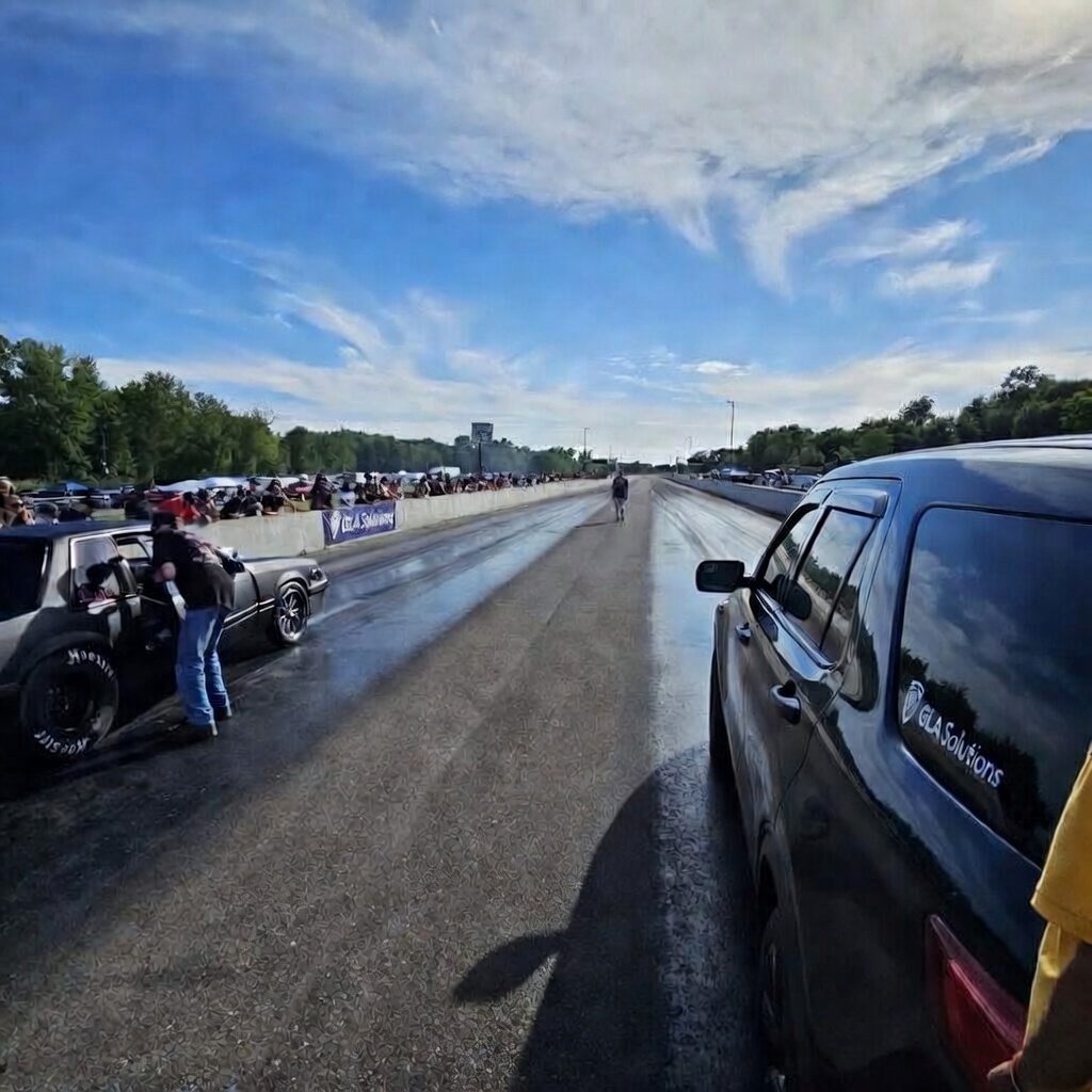 A drag racing event with cars on the side of the track and spectators watching from the stands under a clear blue sky.