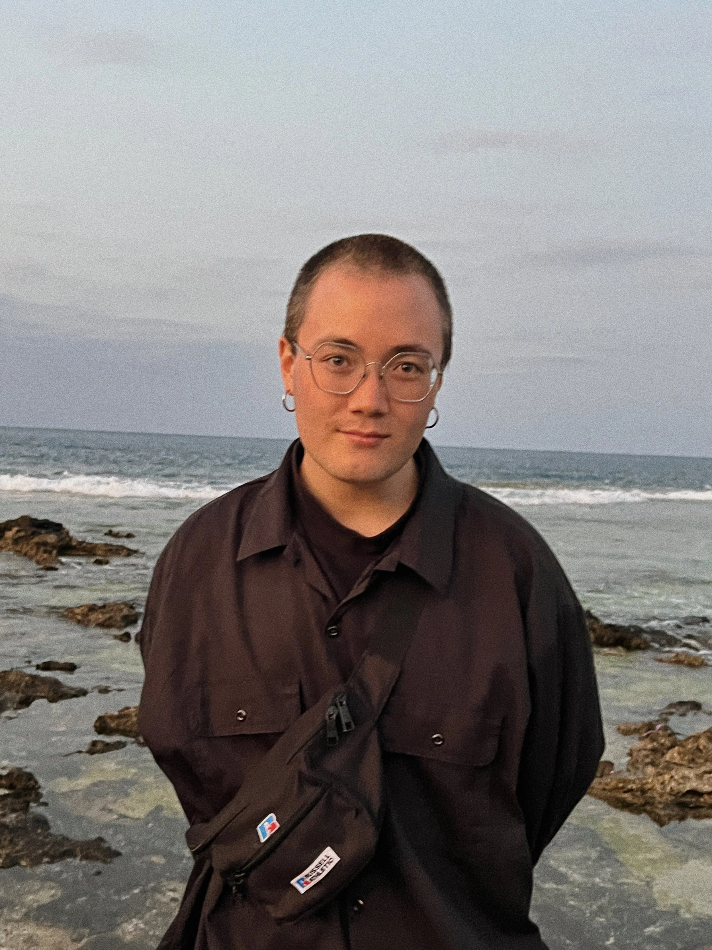 A person with short hair, wearing glasses and earrings, dressed in black, standing near a rocky beach with waves and cloudy sky in the background.
