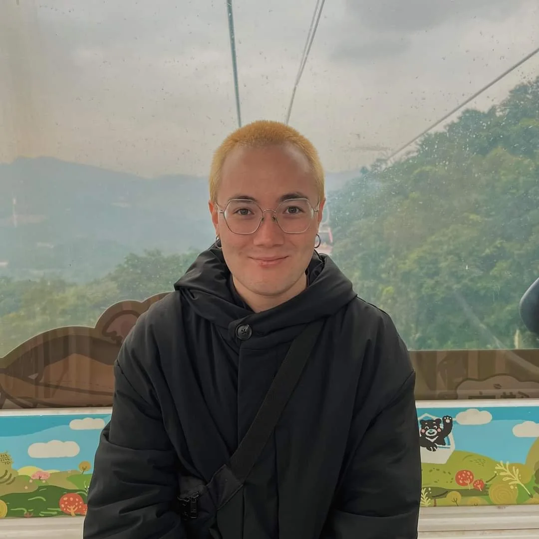 A person with short blond hair, glasses, and earrings, wearing a black coat, smiling in front of a glass window with scenic outdoor view.