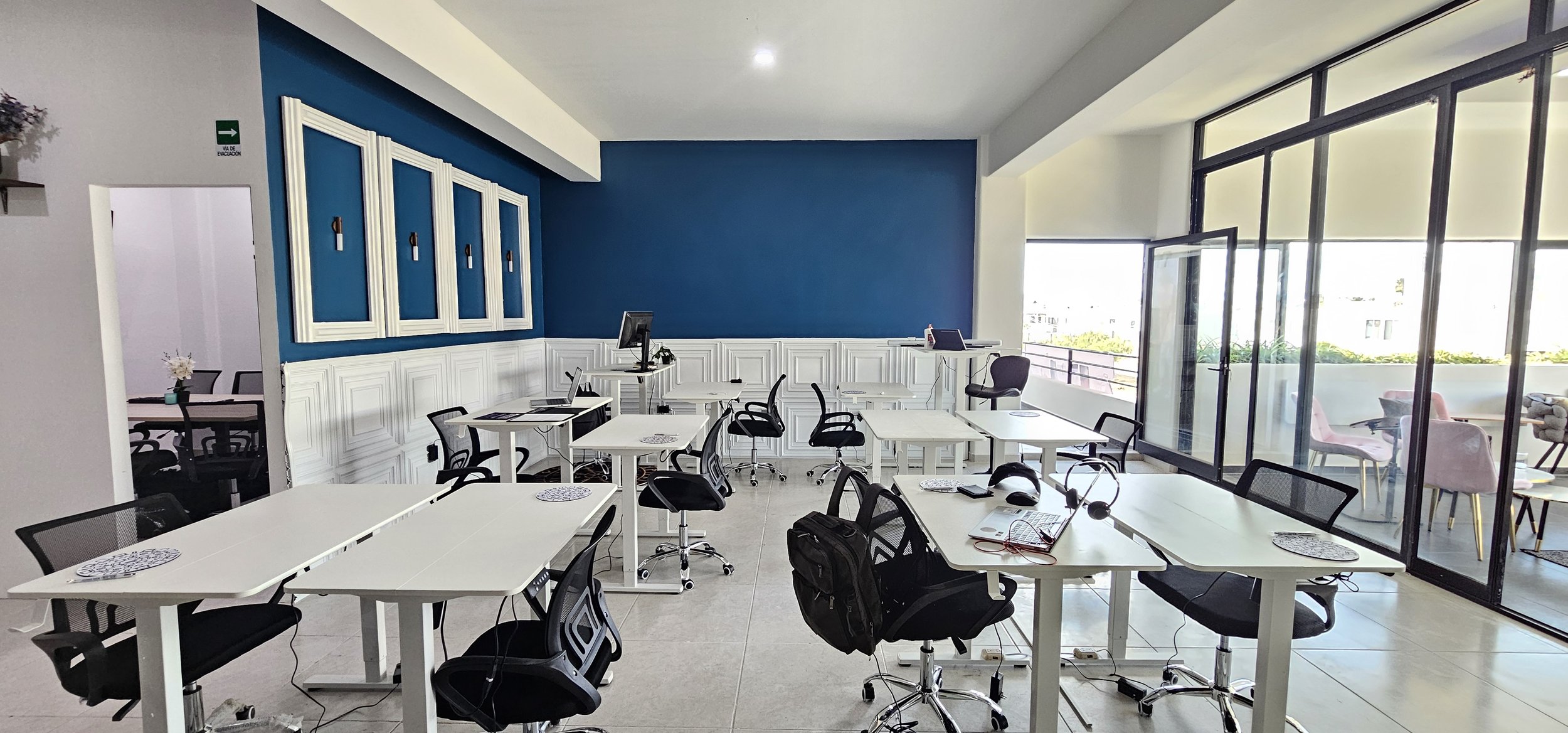 Sala de trabajo con escritorios blancos, sillas negras y una pared azul en el fondo, con ventanas grandes que dejan entrar luz natural, y una puerta hacia otra habitación en la izquierda.