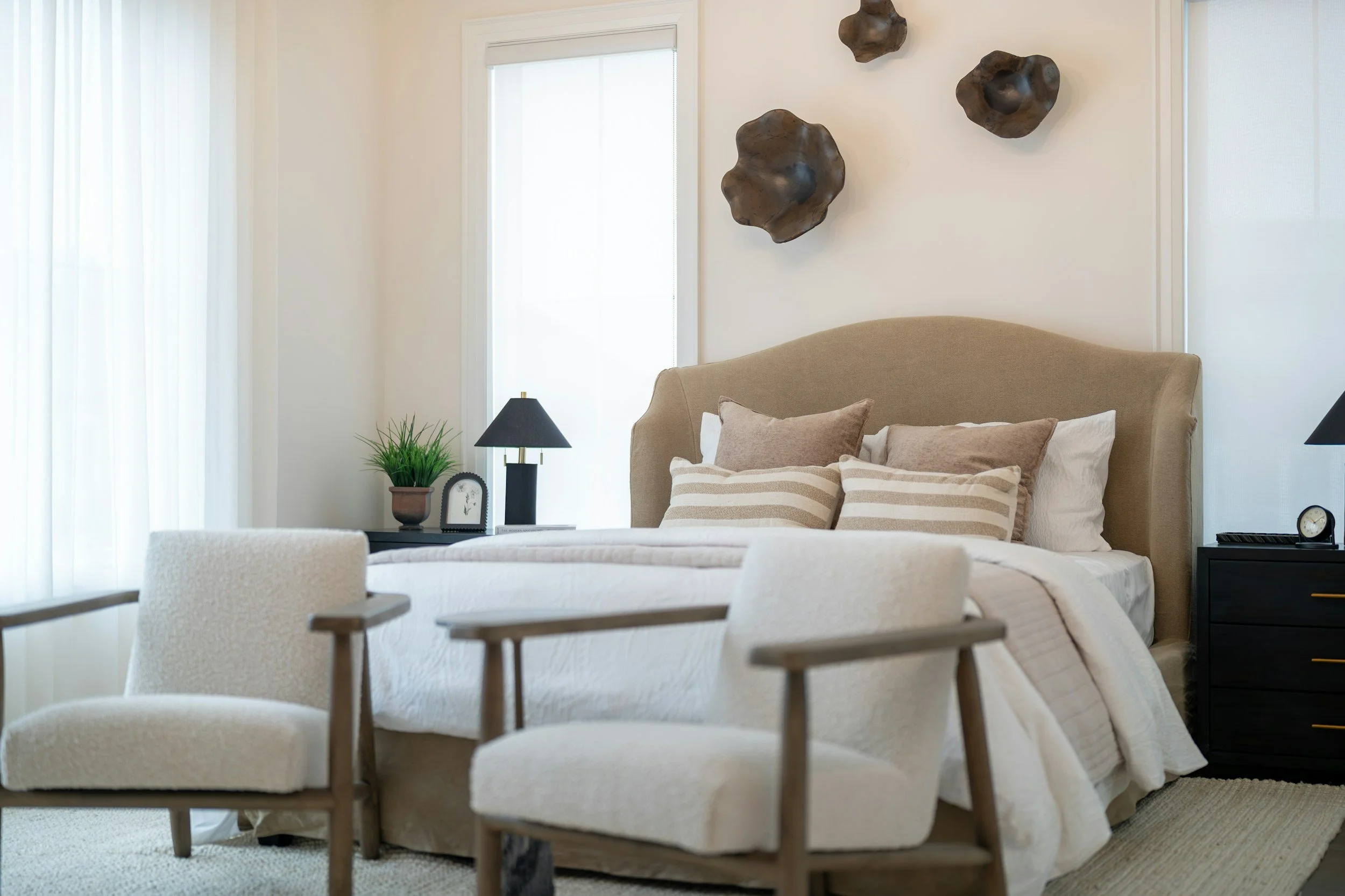 A cozy bedroom with a beige upholstered bed, soft pillows, and white bedding, flanked by black nightstands with lamps, a potted plant, and decorative wall art above.