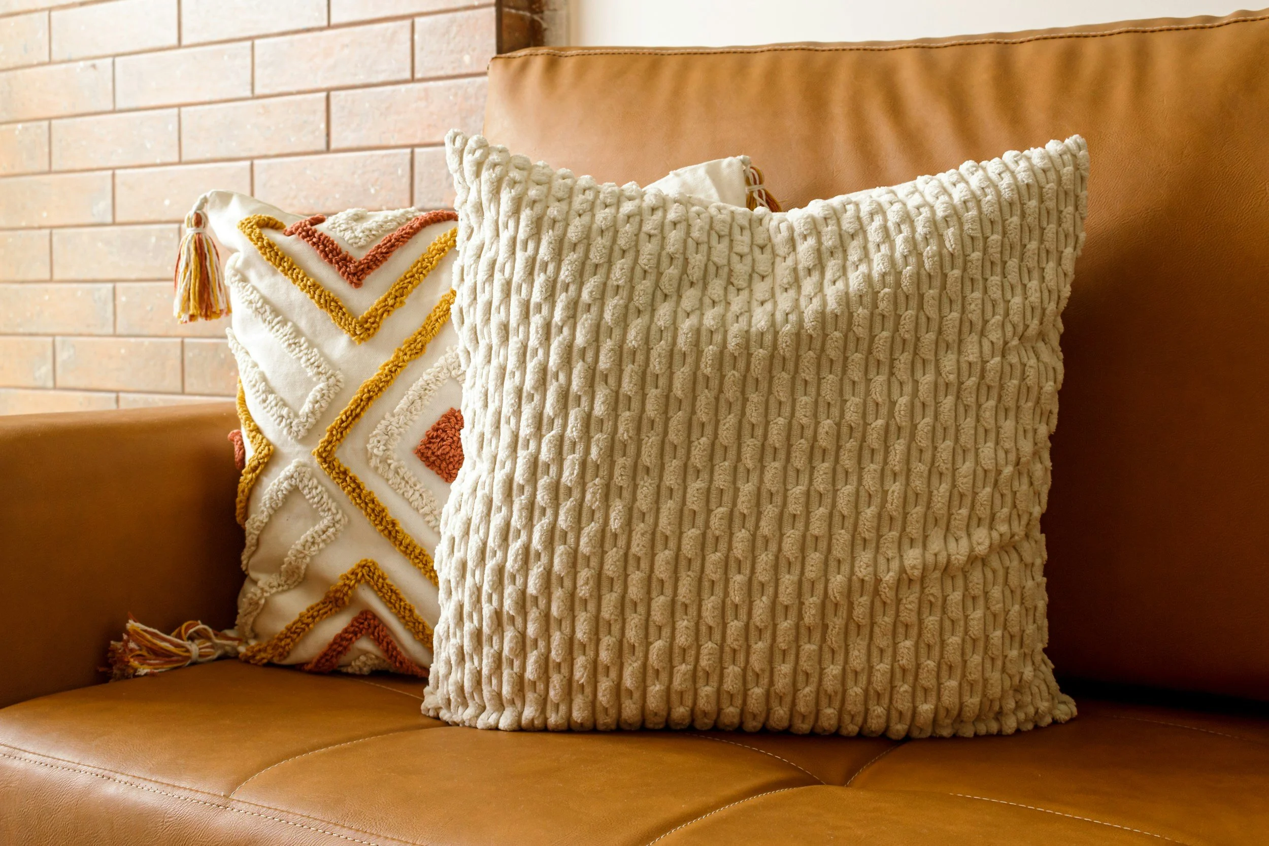 Two decorative throw pillows on a leather sofa, with a brick wall in the background. One pillow has a textured, knitted pattern in beige, and the other features embroidered chevron designs with tassels.