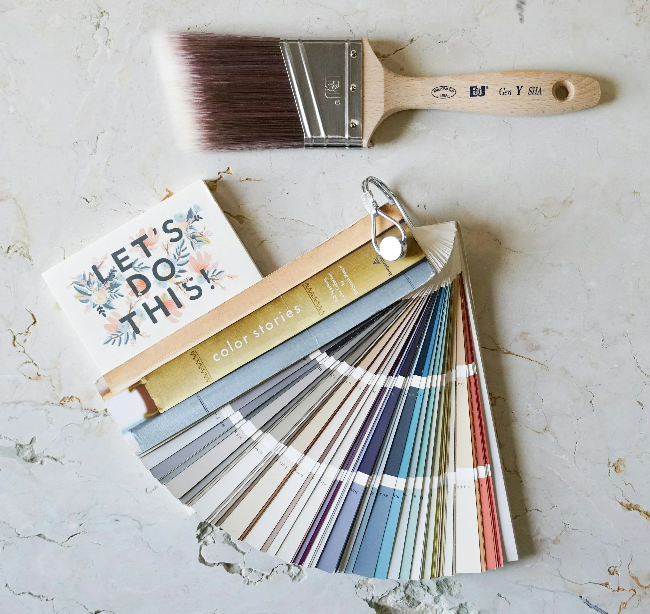 A color swatch fan deck, a paintbrush, a greeting card with flowers and the words "LET'S DO THIS!", and a small metal line clip on a marble surface.