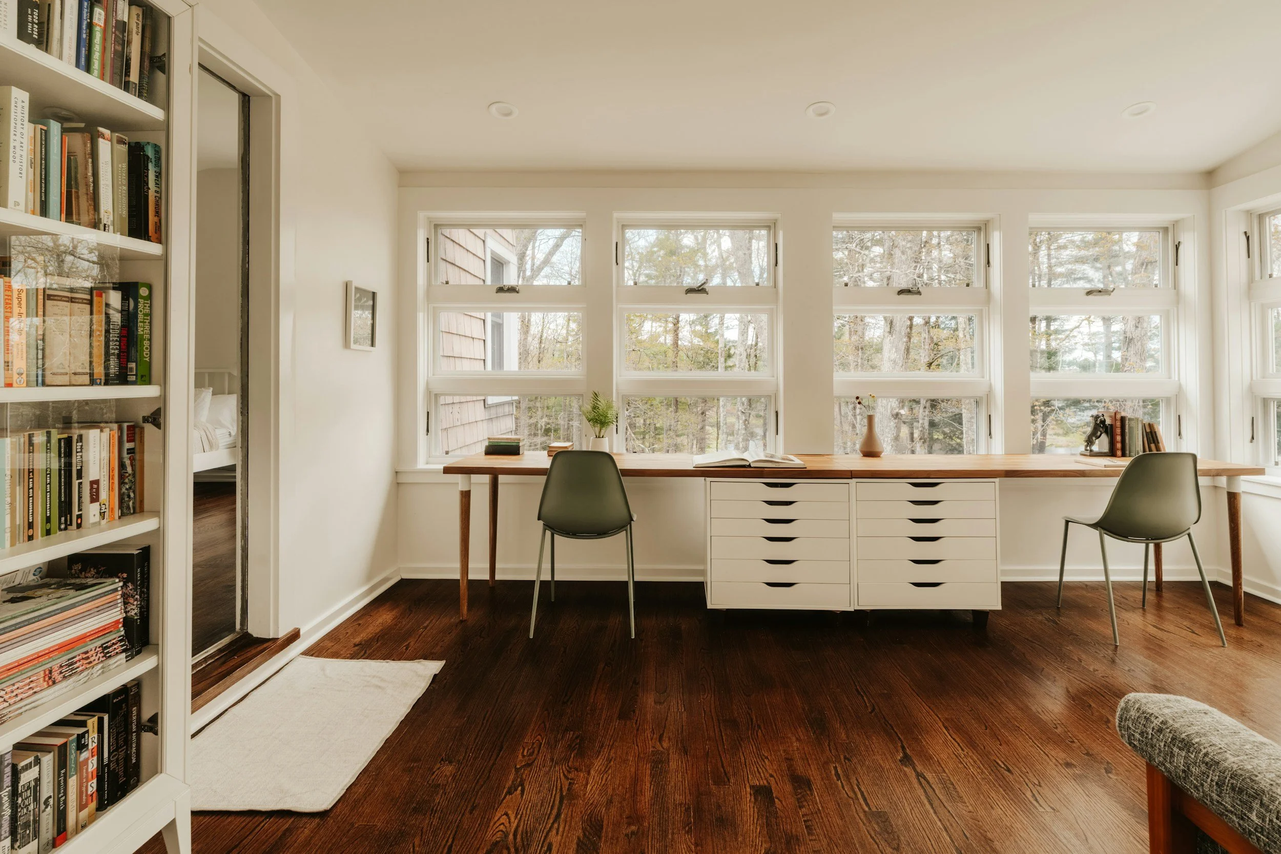 A bright room with large windows overlooking trees, a desk with a plant and books, dark wooden floor, white walls, and a nearby bookshelf.