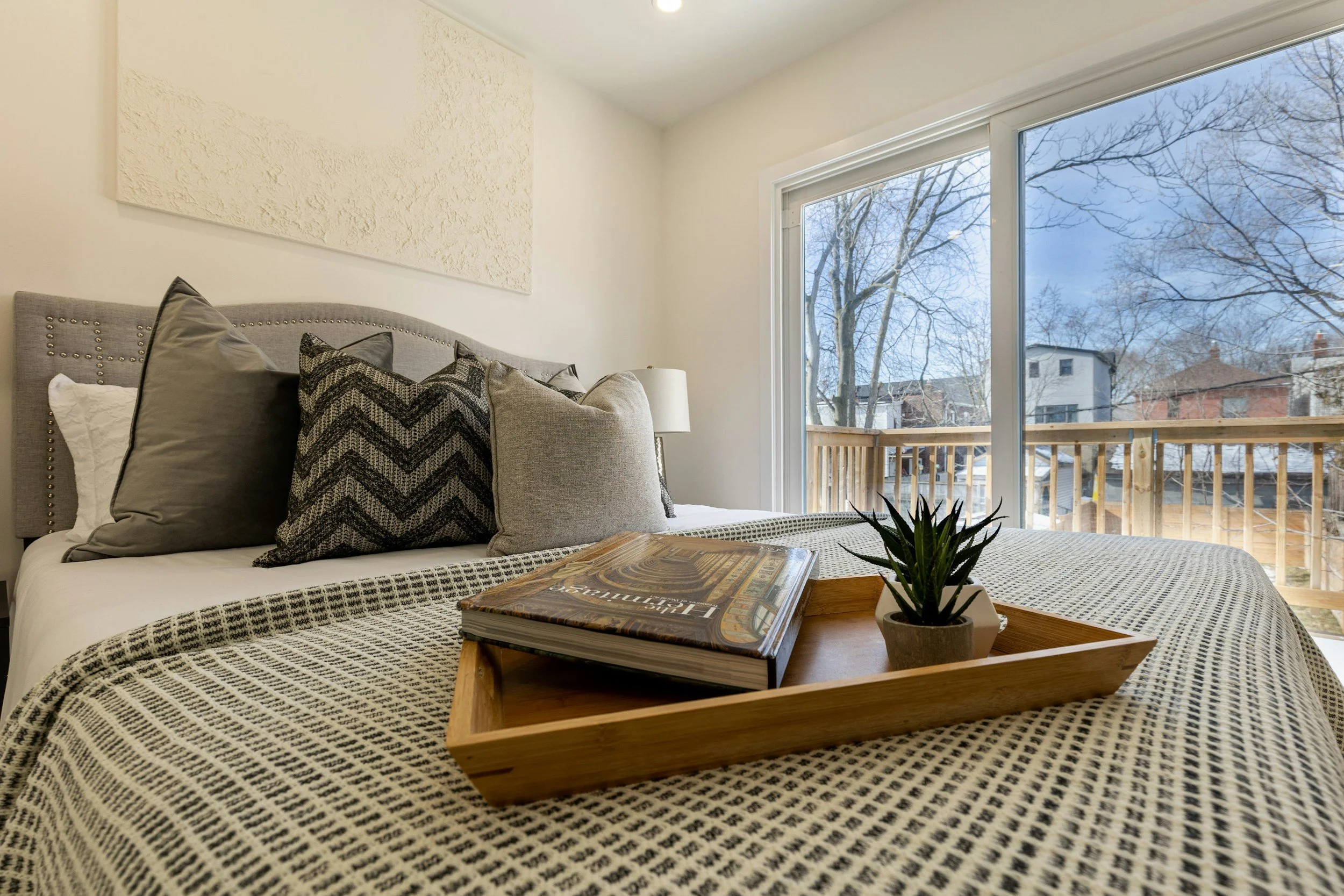 A bedroom with a large window showing a snowy outdoor scene with trees and houses. The bed has a gray upholstered headboard and is decorated with gray, black, and white pillows. On the bed is a wooden tray with a book and a potted plant. A white textured wall art piece hangs above the headboard, and a small white table lamp is beside the bed.