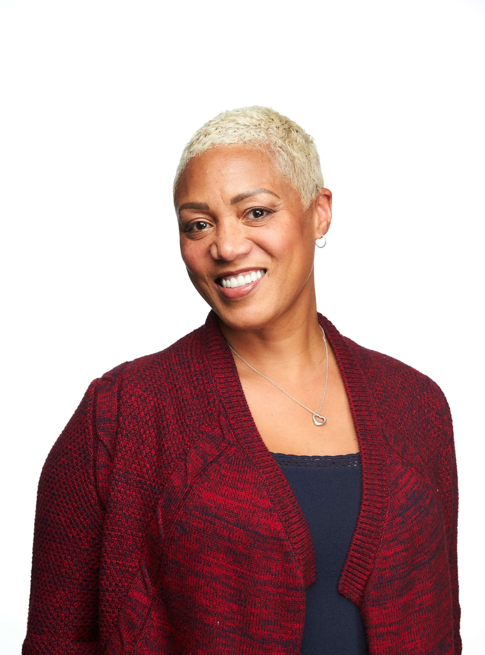 A woman with short, bleach blonde hair smiling, wearing a red blazer over a navy top with a silver heart necklace, set against a white background.