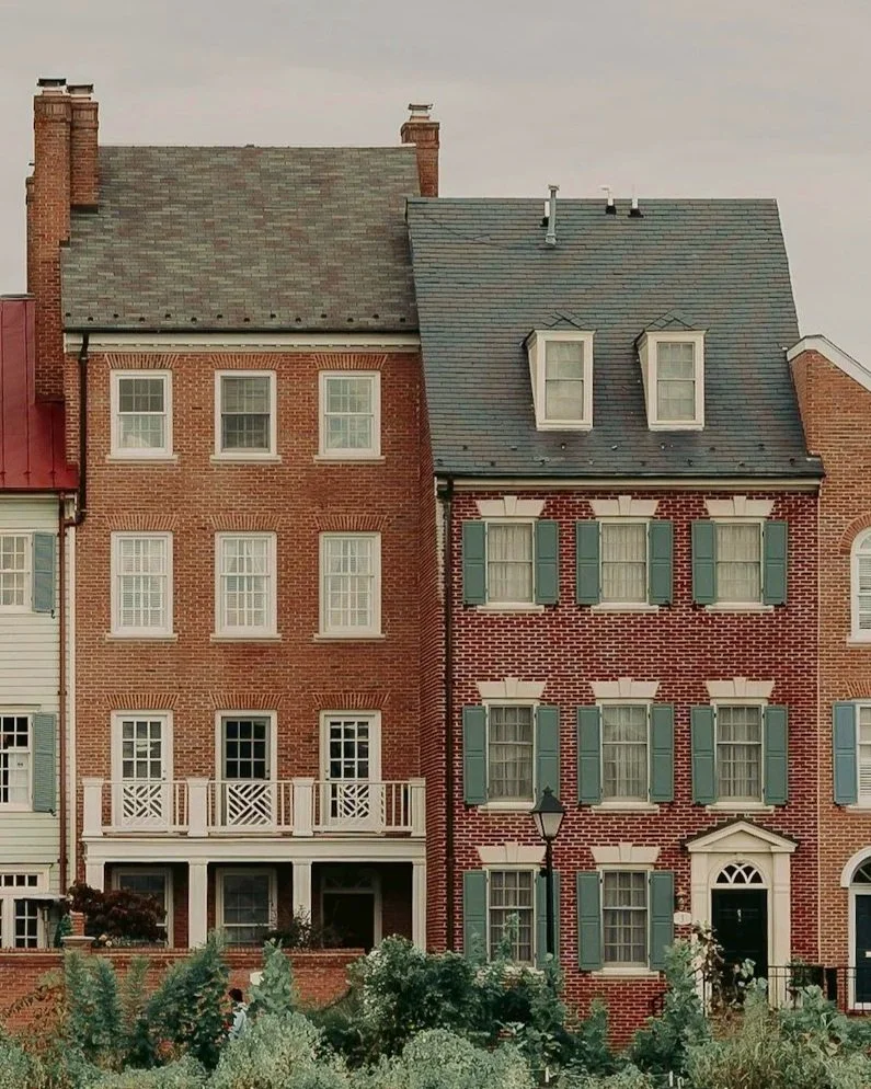 Three brick townhouses with different architectural styles and window arrangements, surrounded by greenery.