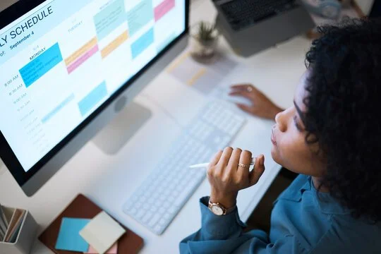 A woman looking at a computer screen displaying a weekly schedule or calendar with color-coded blocks.