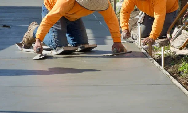 Two construction workers in orange shirts and jeans smoothing wet concrete with trowels.