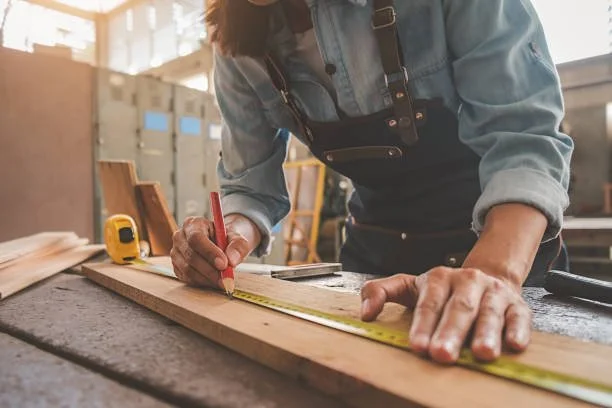 A person working in a woodworking shop, measuring a piece of wood with a tape measure.