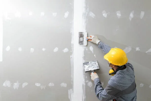 A construction worker in a yellow helmet is installing drywall on a wall in a building under construction.