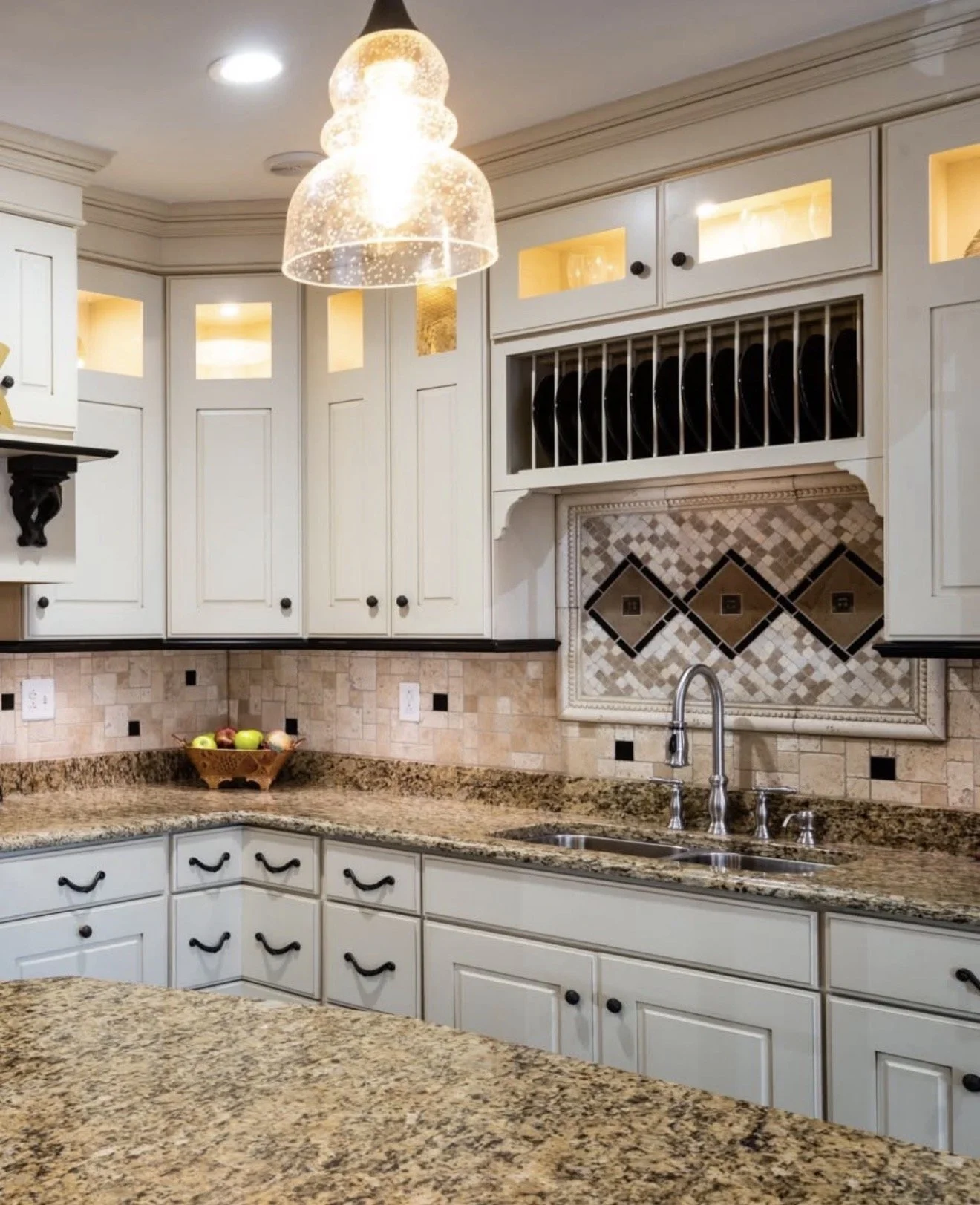 Kitchen with white cabinets, granite countertop, a basket of apples, tiled backsplash, double sink, and a decorative range hood with plate rack. Overhead lighting and a hanging glass pendant light.