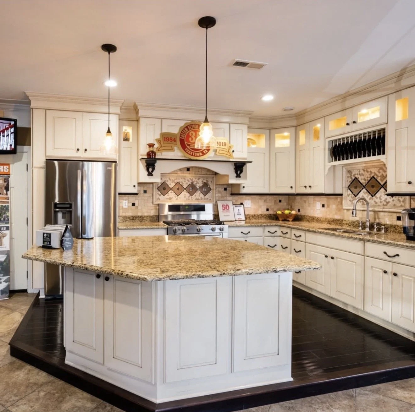 by Custom Design Works Inc. | kitchen with white cabinets, granite countertops, a central island, stainless steel refrigerator, stove, and a double sink. Decor includes a bowls of fruit and framed awards and decorative items on top of the cabinets.