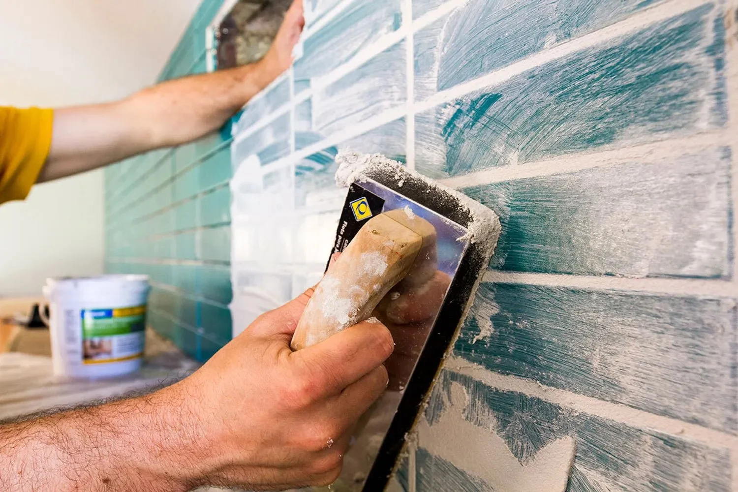 Person scraping tiles on a wall with a trowel, applying grout during wall tile installation.